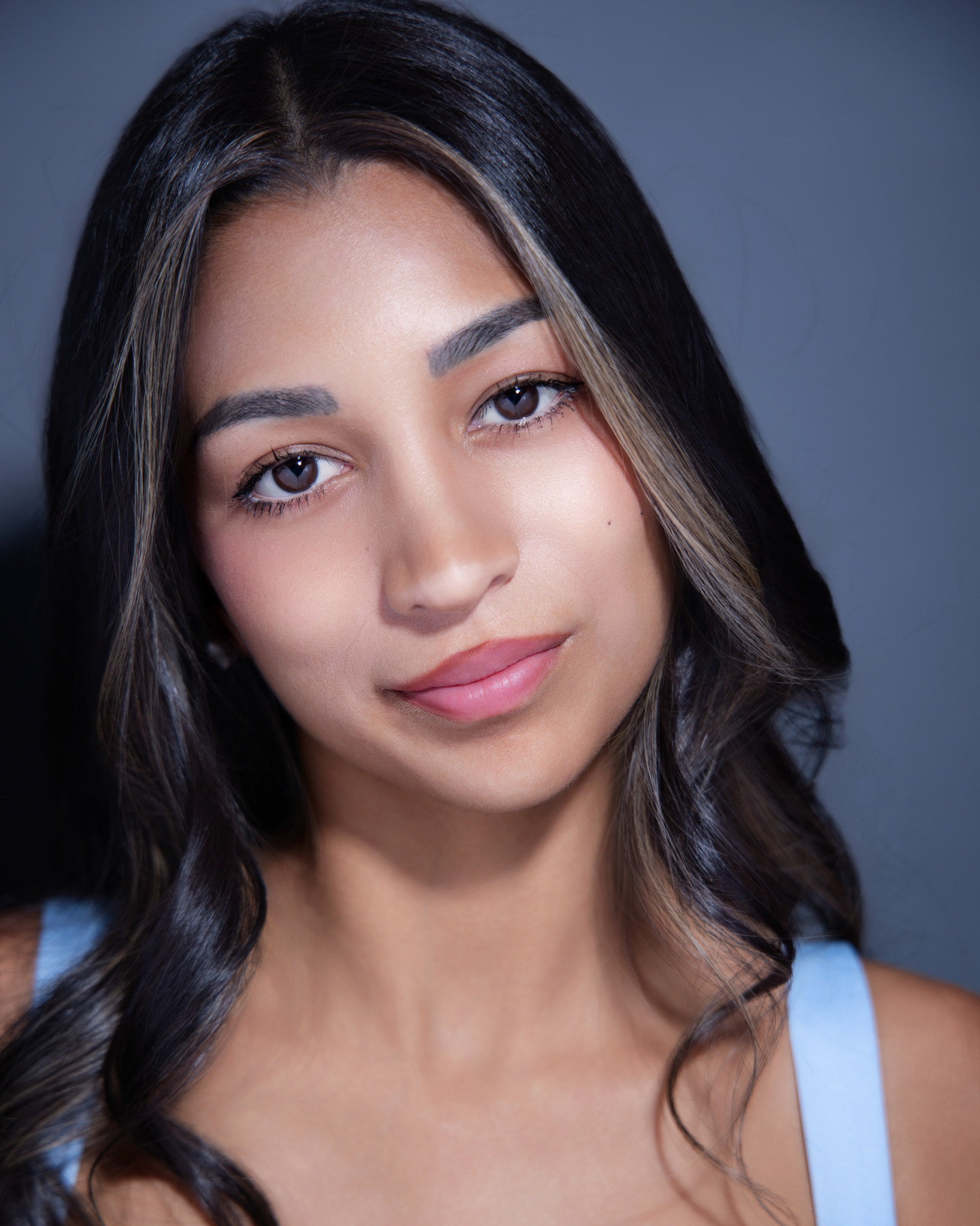 Close-up portrait of a woman with long dark hair in a light blue top against a gray backdrop