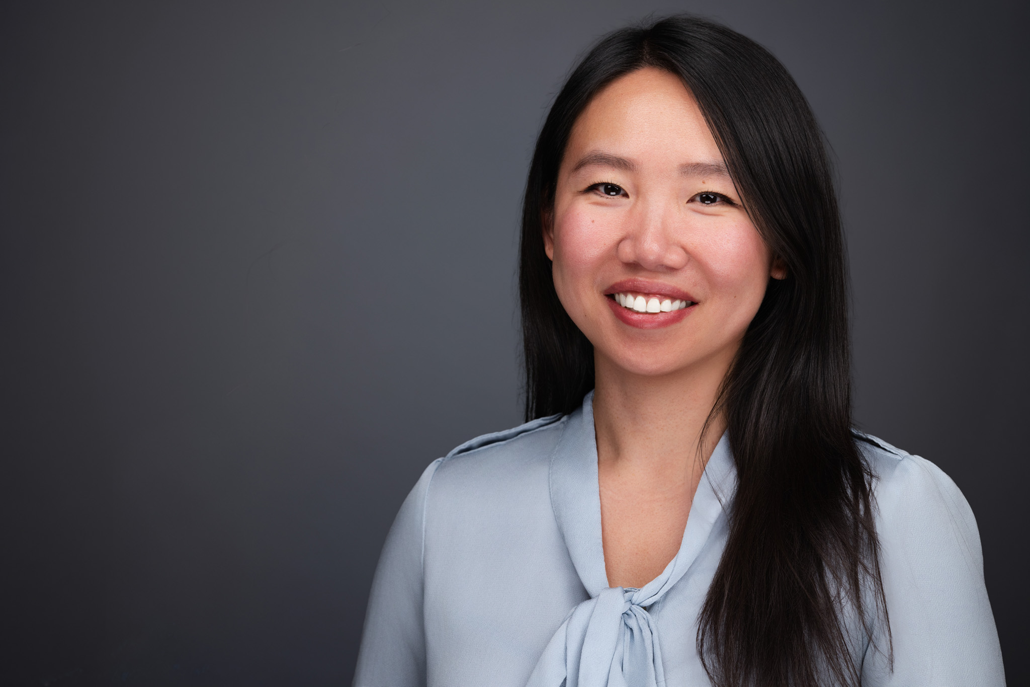 Woman with long dark hair in a light blue top, studio headshot against a dark gray backdrop