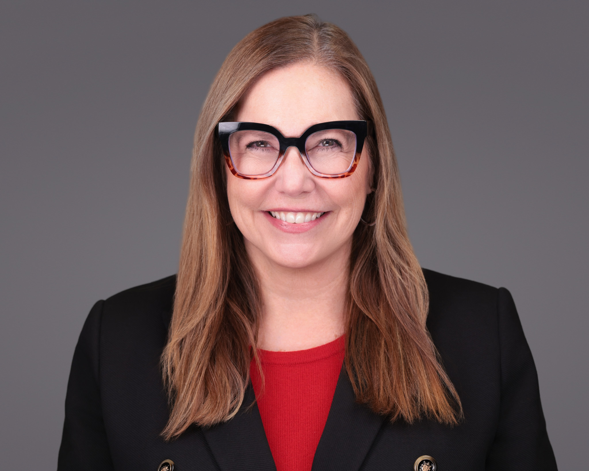 Woman with glasses and long hair in a black blazer and red top, studio headshot against a dark gray backdrop