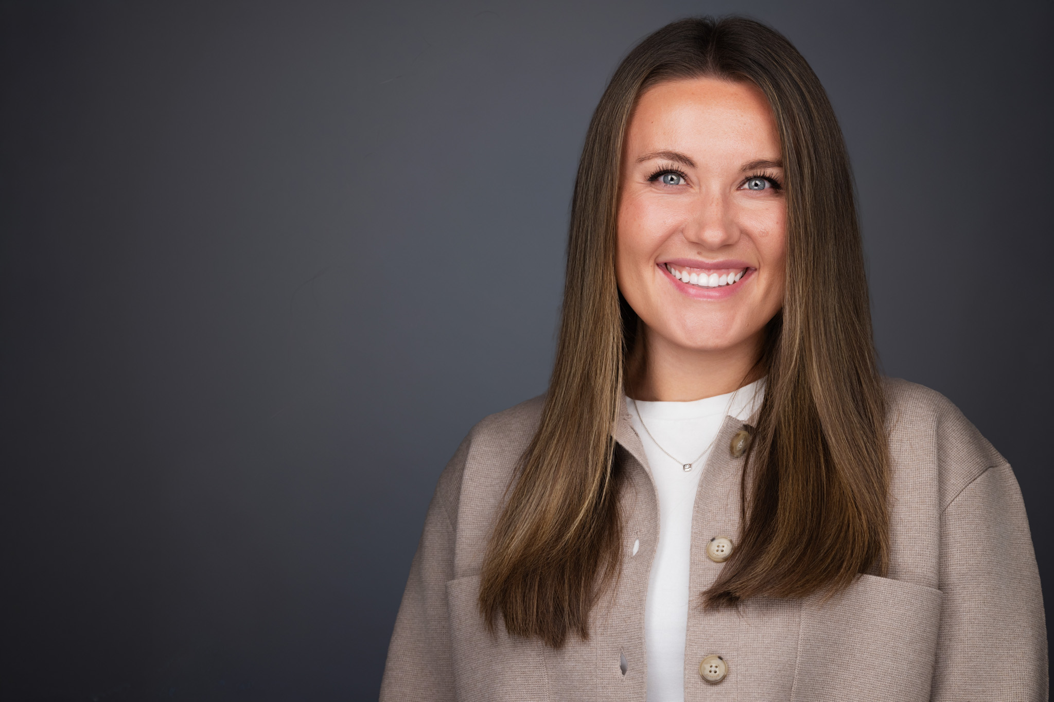 Smiling woman with long brown hair in a cream top, studio headshot against a dark gray backdrop
