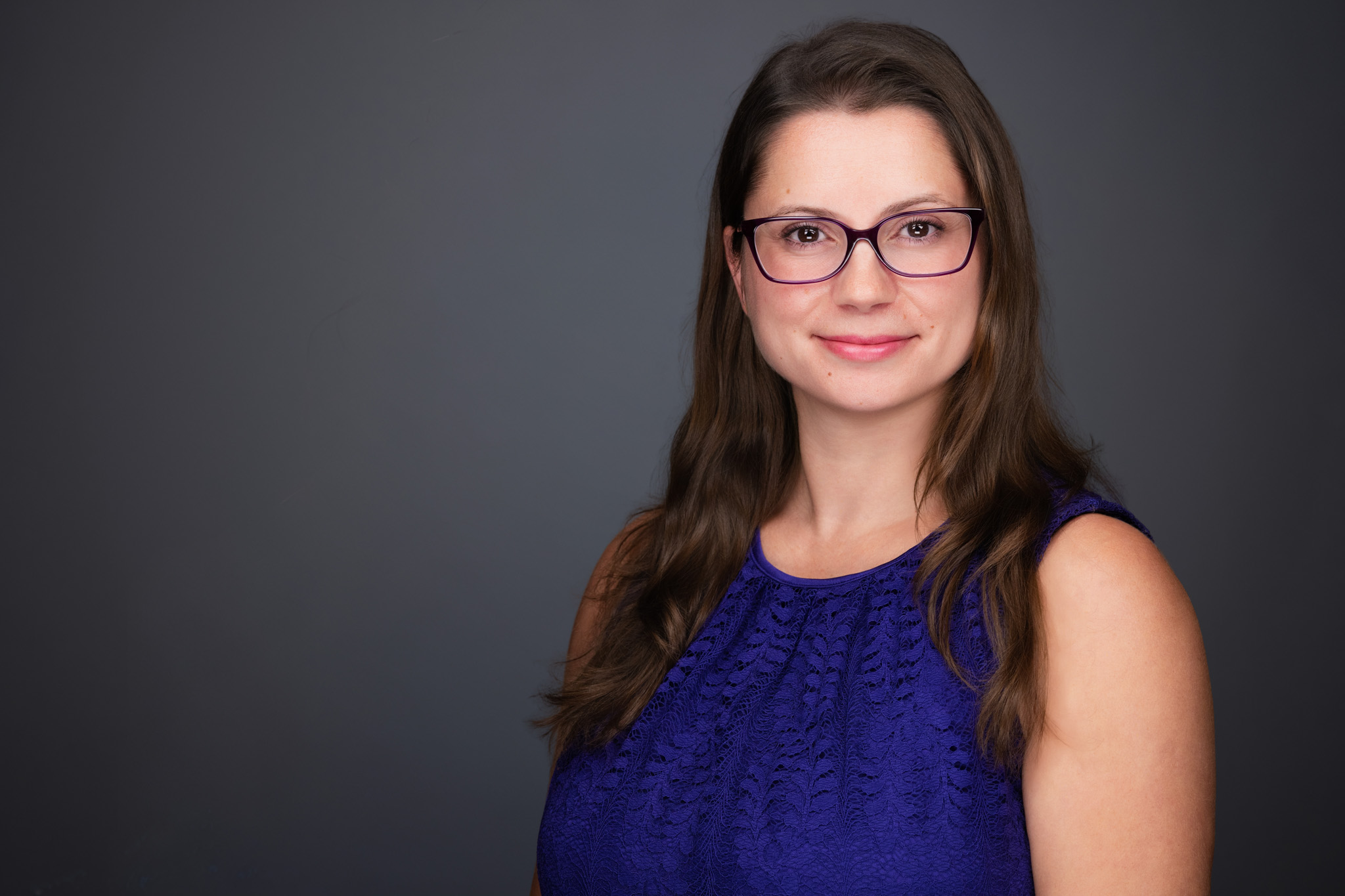 Smiling woman with long brown hair and glasses in a blue top, studio headshot against a dark gray backdrop