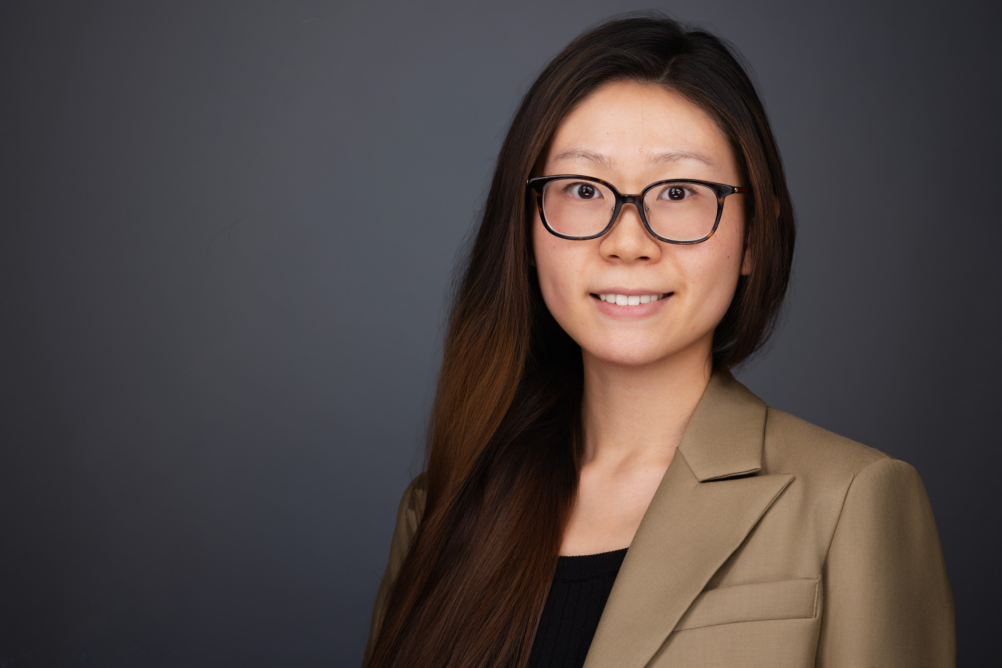 Woman with glasses and long dark hair in a tan blazer, studio headshot against a dark gray backdrop