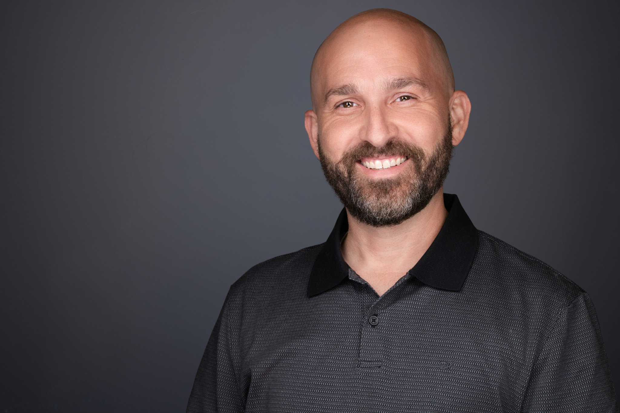 Man with a shaved head and beard in a dark shirt, studio headshot against a dark gray backdrop