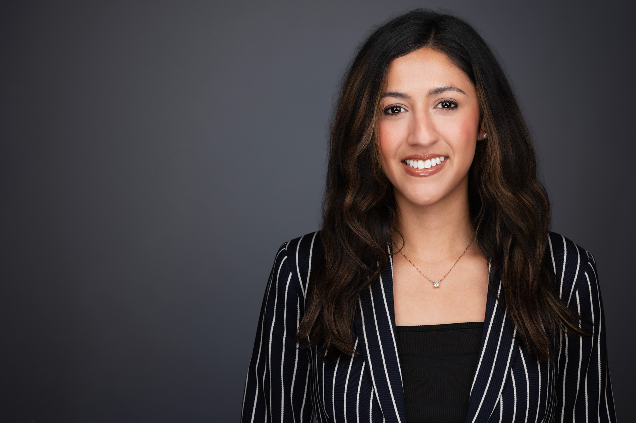 Smiling woman with long dark hair in a striped blazer, studio headshot against a dark gray backdrop