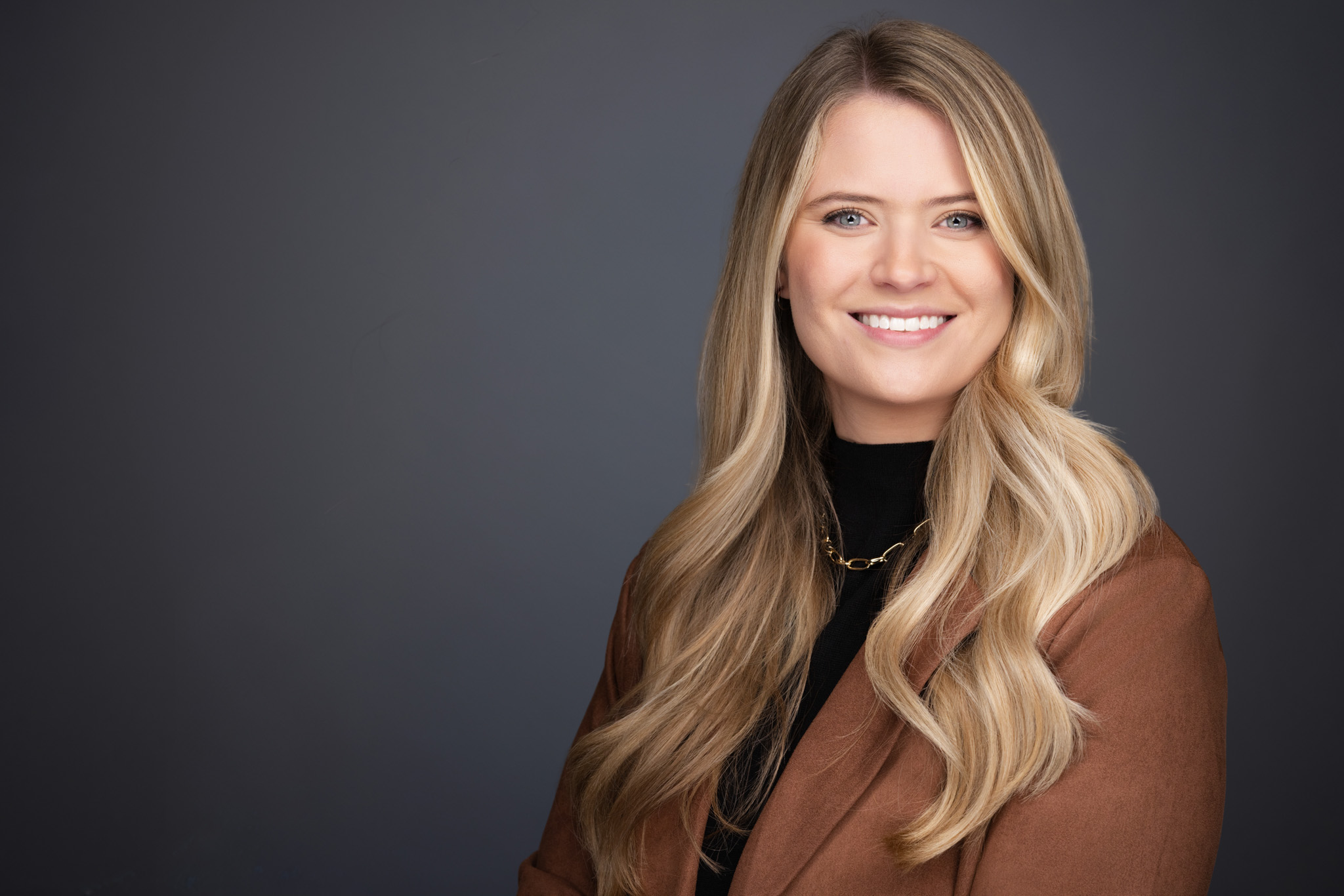 Smiling woman with long blonde hair in a brown jacket, studio headshot against a dark gray backdrop