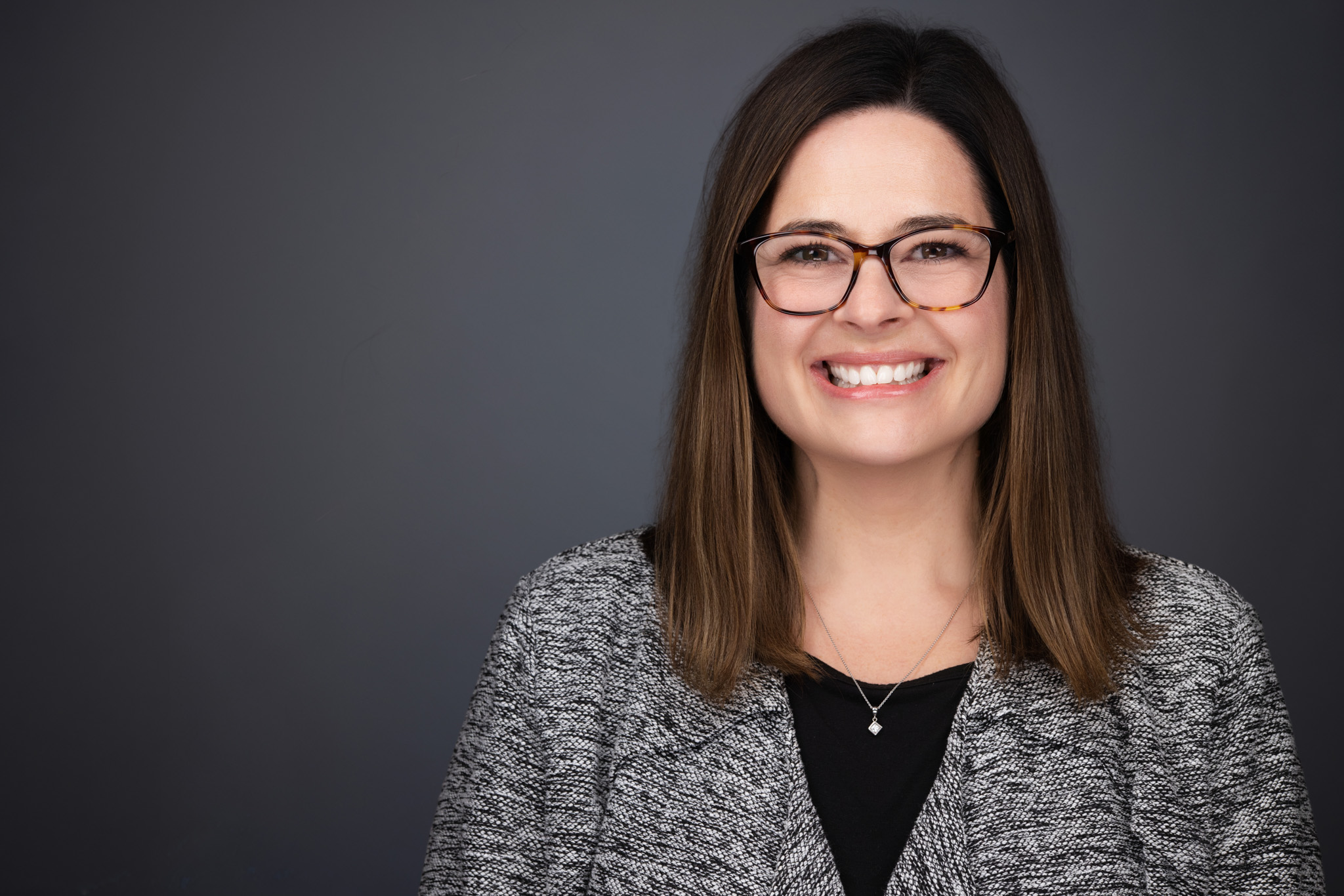 Woman with glasses and dark hair in a gray cardigan, studio headshot against a dark gray backdrop