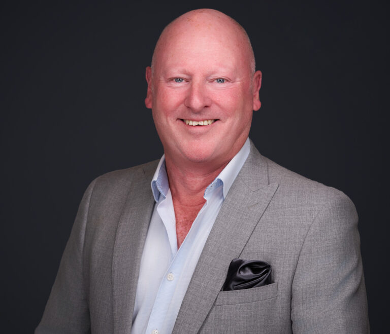 Professional headshot of a smiling bald man wearing a gray blazer and light open-collar shirt against a dark background