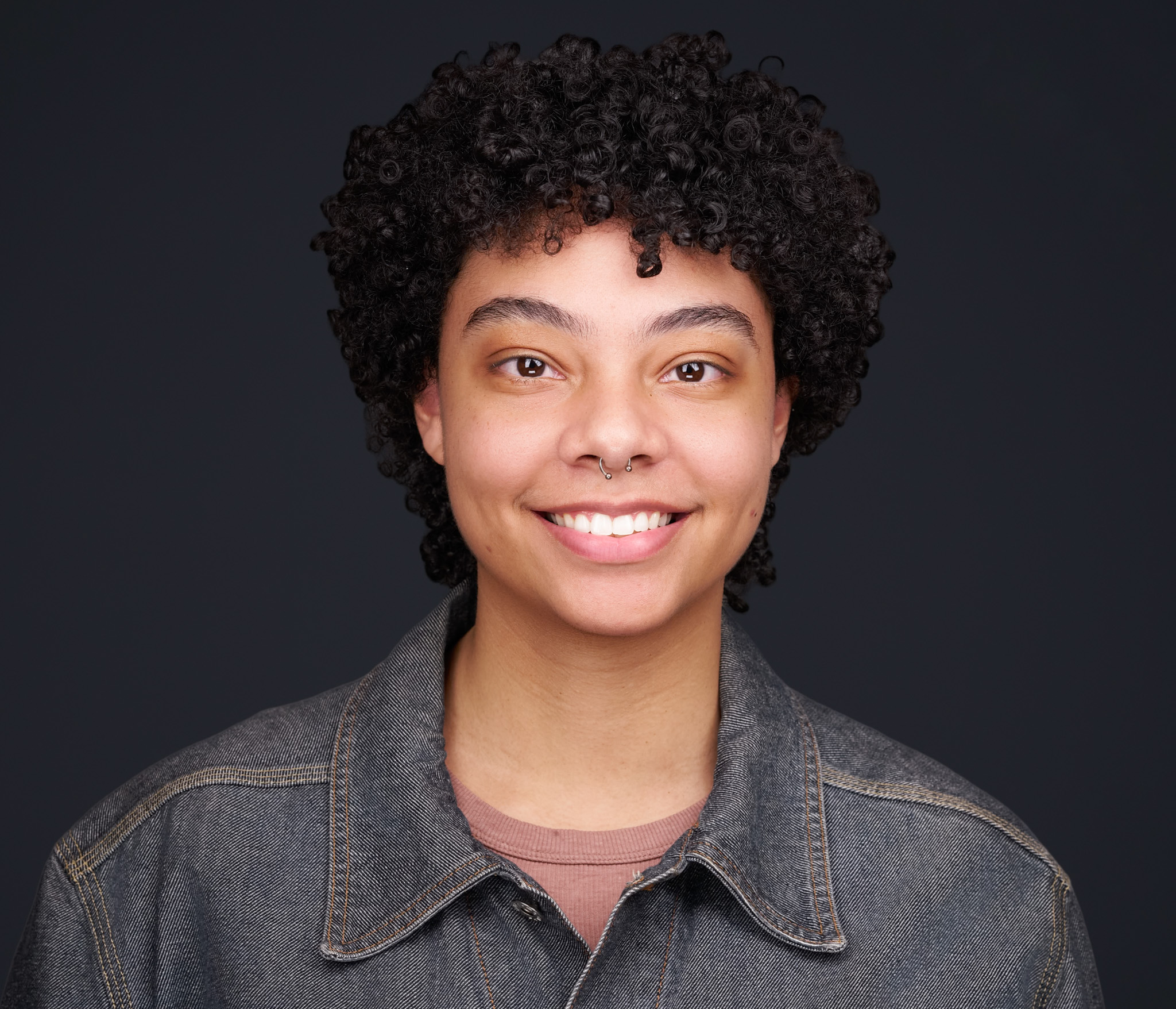 Smiling woman with curly dark hair in a denim jacket, studio headshot against a dark gray backdrop