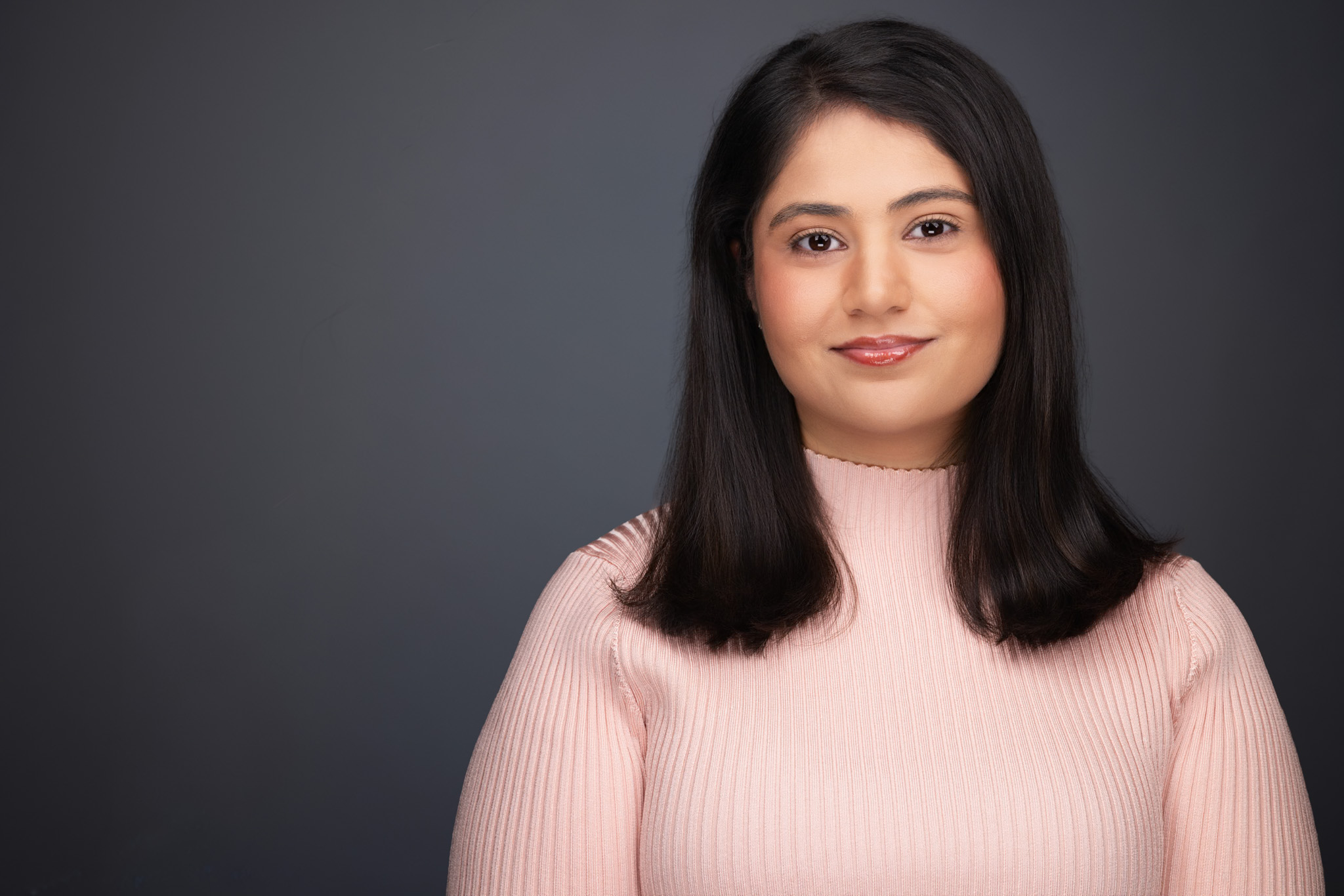 Woman with long dark hair in a pink sweater, studio headshot against a dark gray backdrop