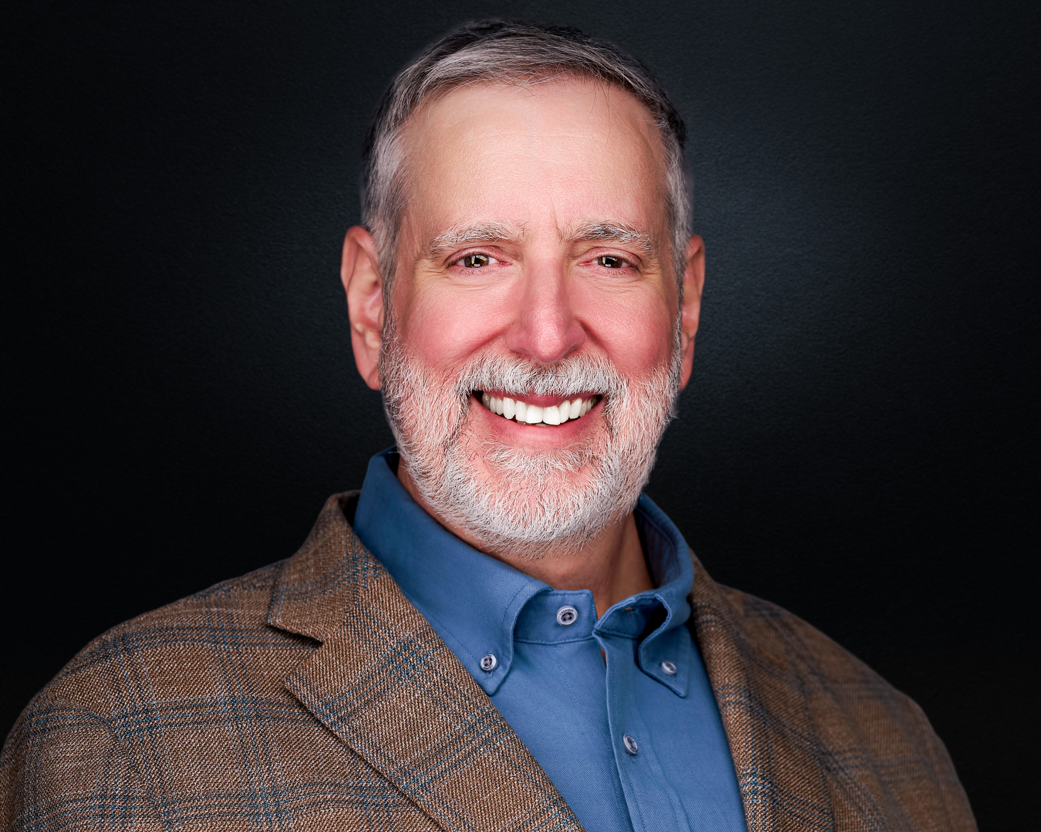 Smiling man with a gray beard in a plaid blazer, studio headshot against a dark gray backdrop