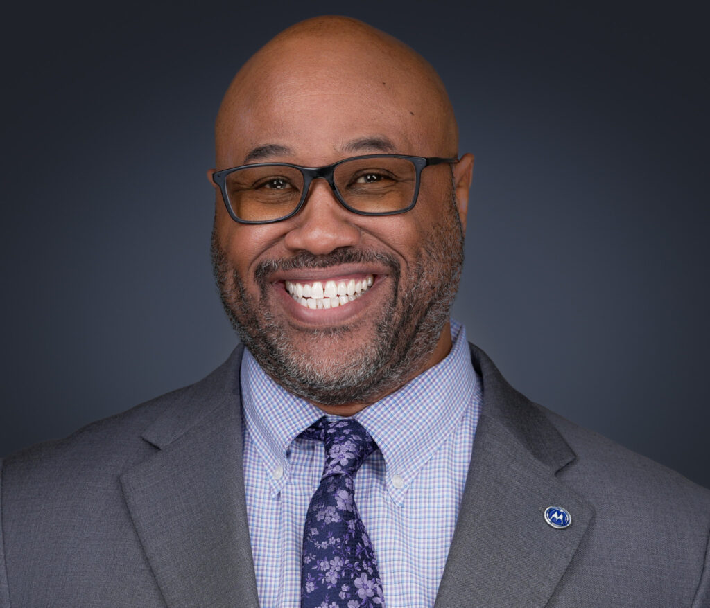 Smiling man with dark-framed glasses wearing a gray suit and purple floral tie, close-up headshot cropped at the top of the head.