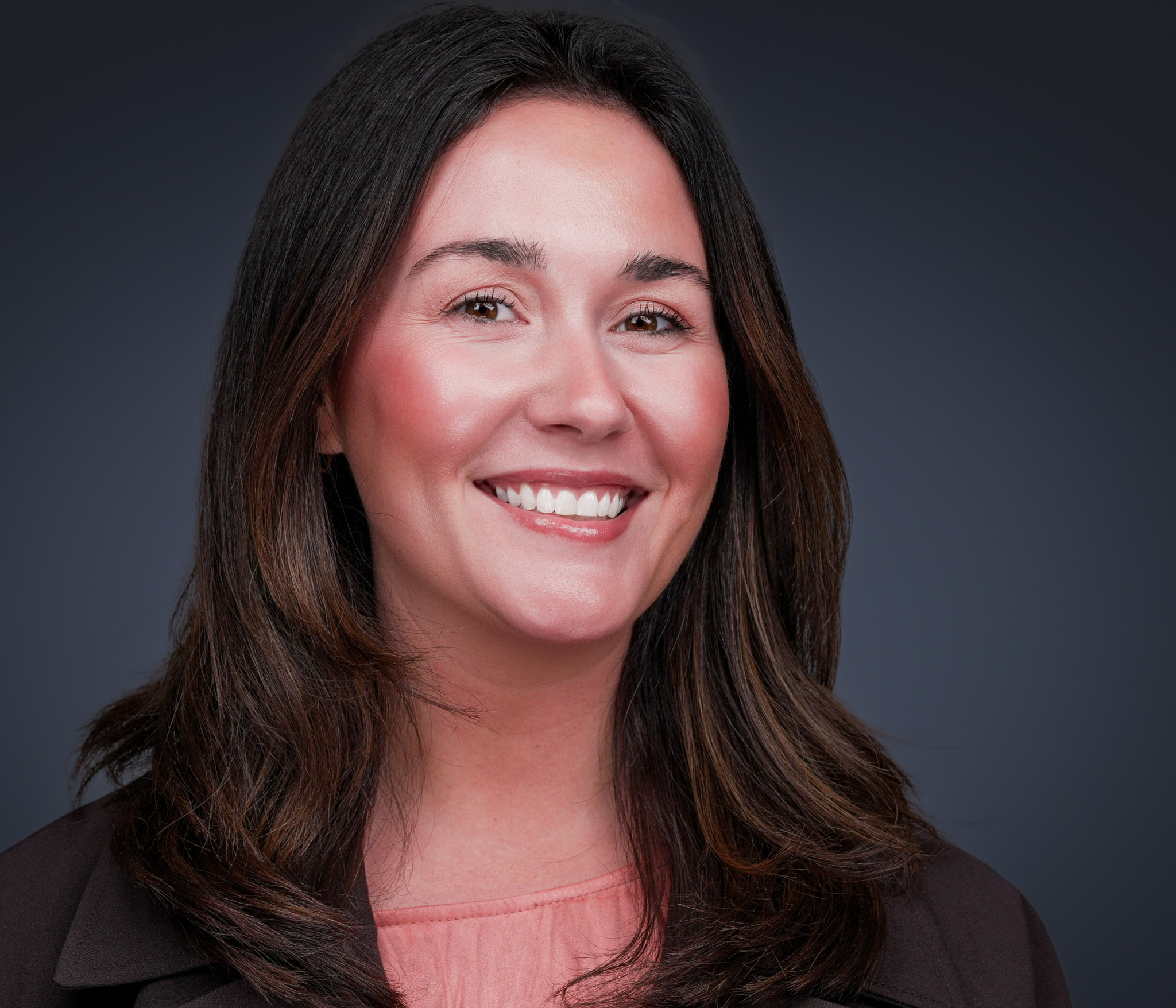 Woman with long dark hair and a warm smile, studio headshot against a gray backdrop