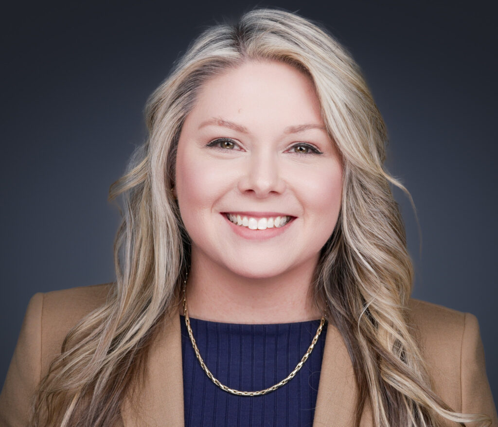 Close-up portrait of a smiling blonde woman wearing a navy top and chain necklace, against a dark gray background.