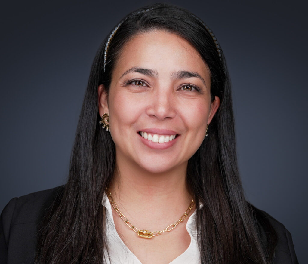 Smiling woman with long dark hair wearing a gold necklace and earrings, white blouse, and black blazer.