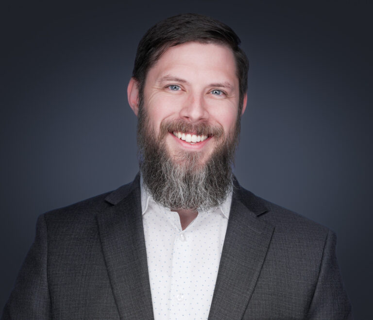 Professional headshot of a smiling bearded man in a dark gray blazer against a dark background