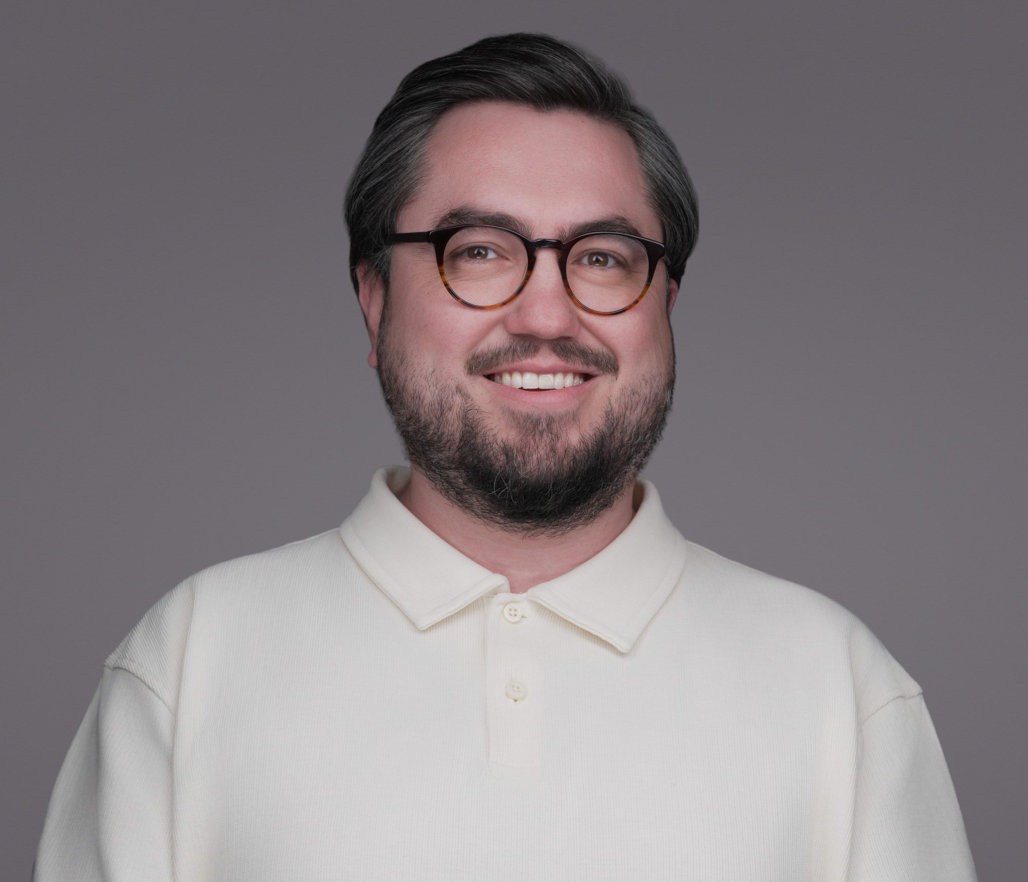 Smiling man with glasses and a beard in a cream sweater, studio headshot against a gray backdrop