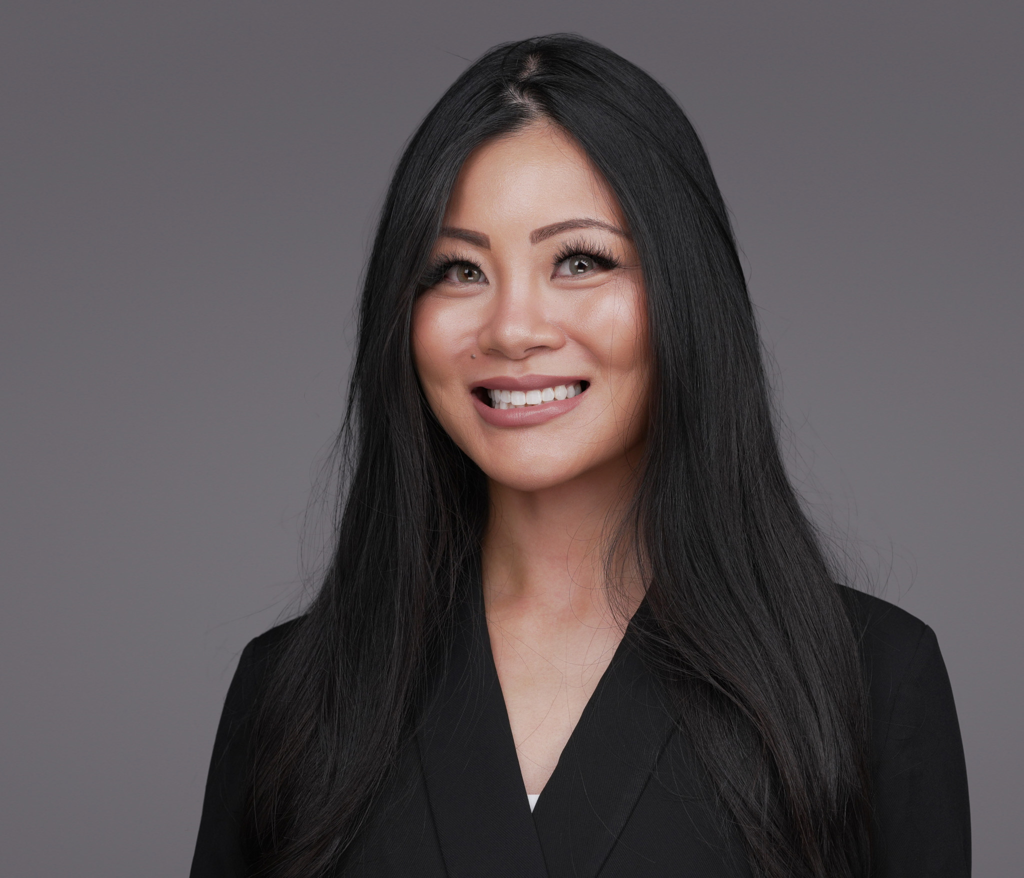 Smiling woman with long black hair in a black blazer, studio headshot against a dark gray backdrop