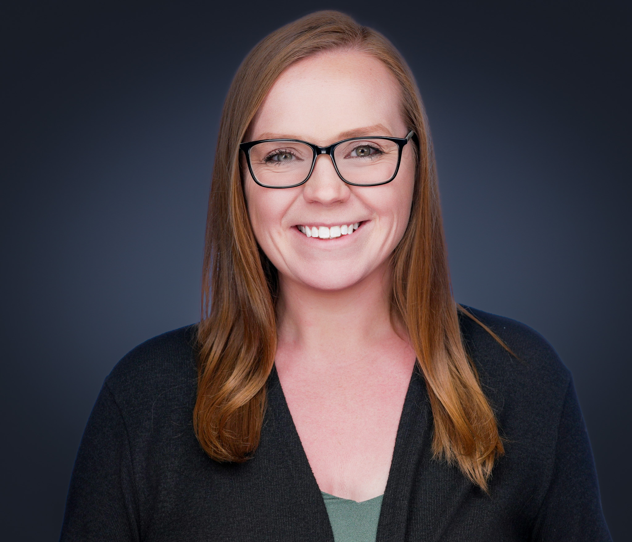 Smiling woman with glasses and long hair in a black blazer, studio headshot against a dark gray backdrop