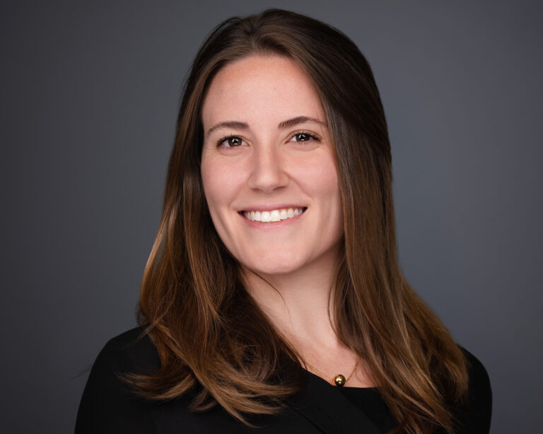 Professional headshot of a smiling woman with long dark brown hair wearing a black top against a gray background