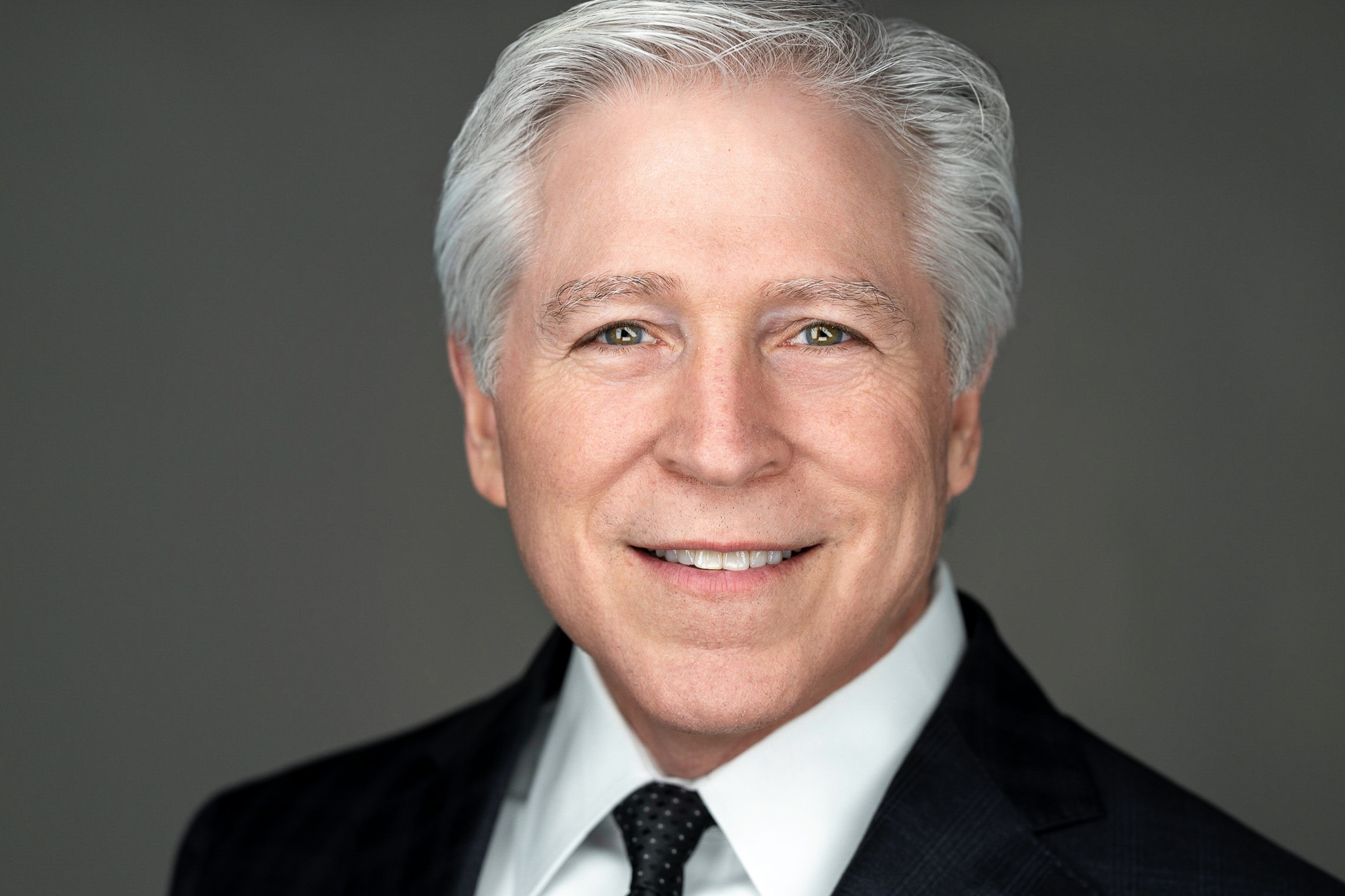 Smiling older man with white hair in a dark suit, studio headshot against a dark gray backdrop