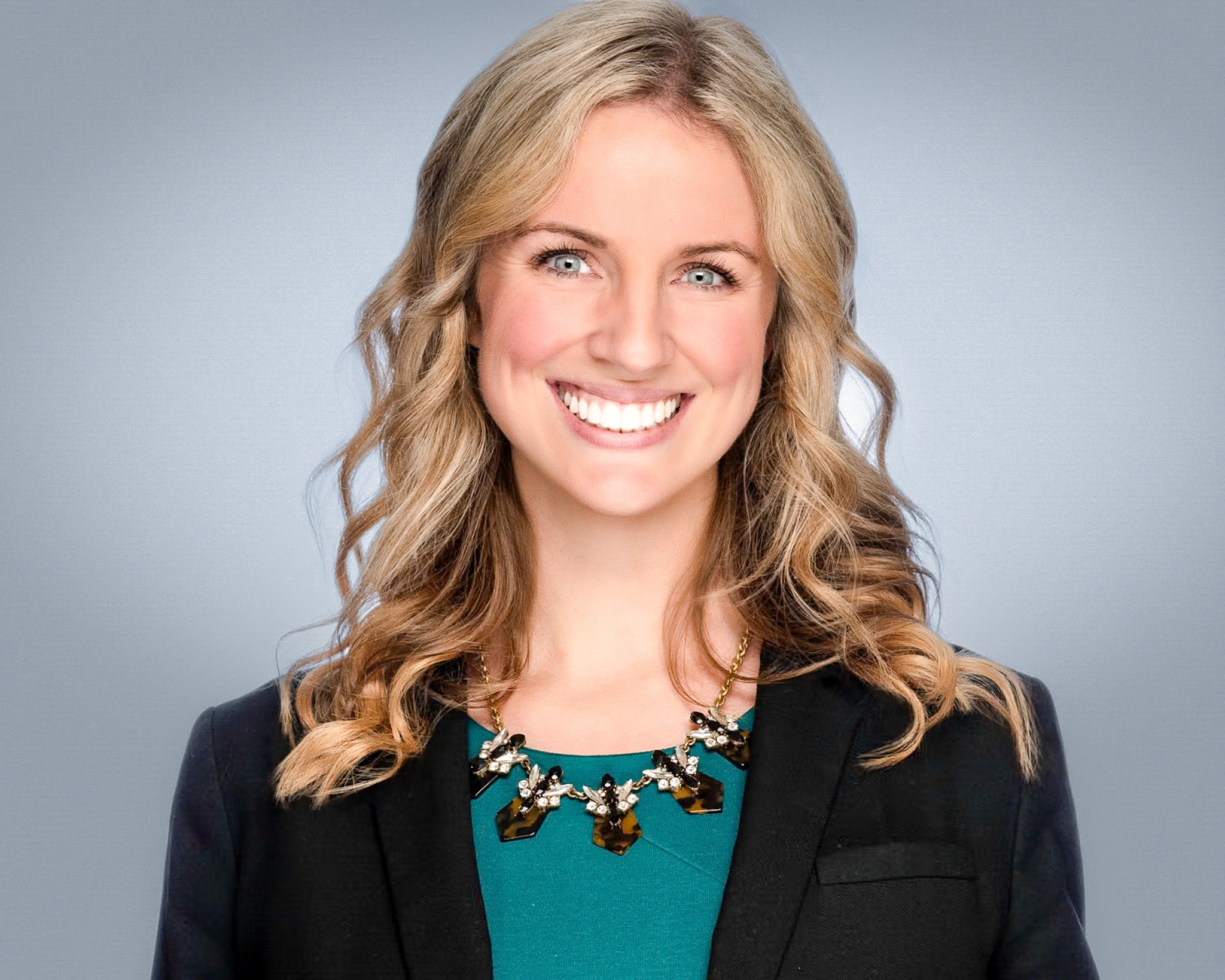 Smiling woman with wavy blonde hair and a teal top, studio headshot against a dark blue backdrop