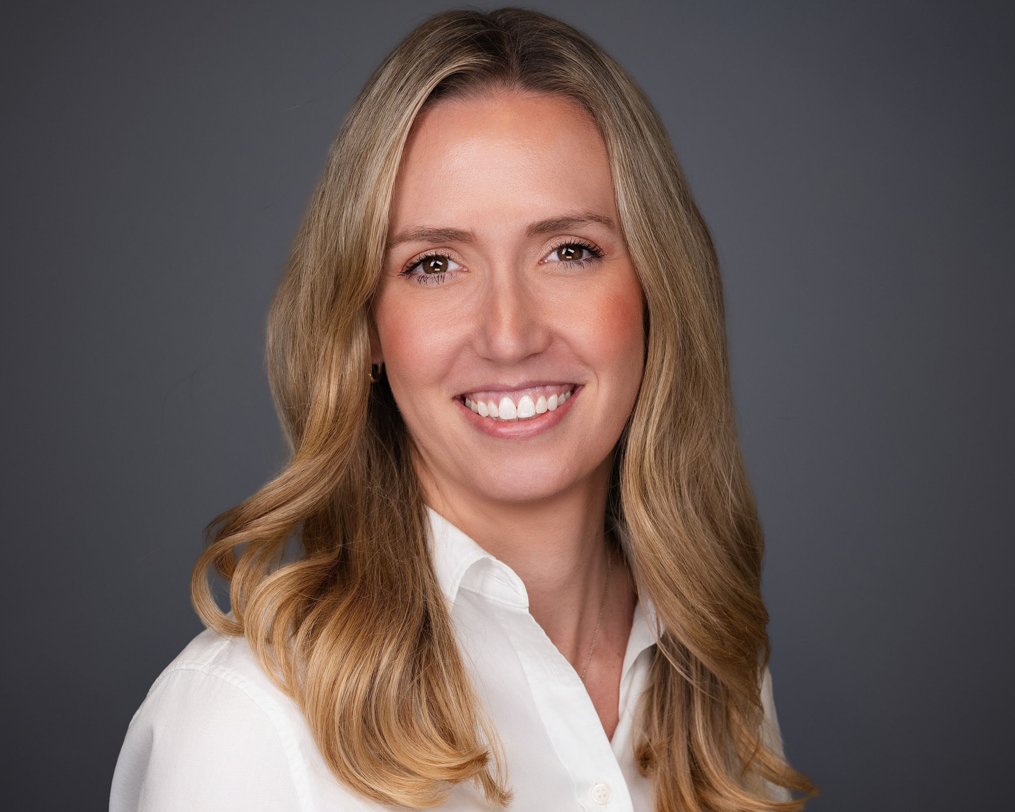 Smiling woman with long blonde hair in a white blouse, studio headshot against a gray backdrop
