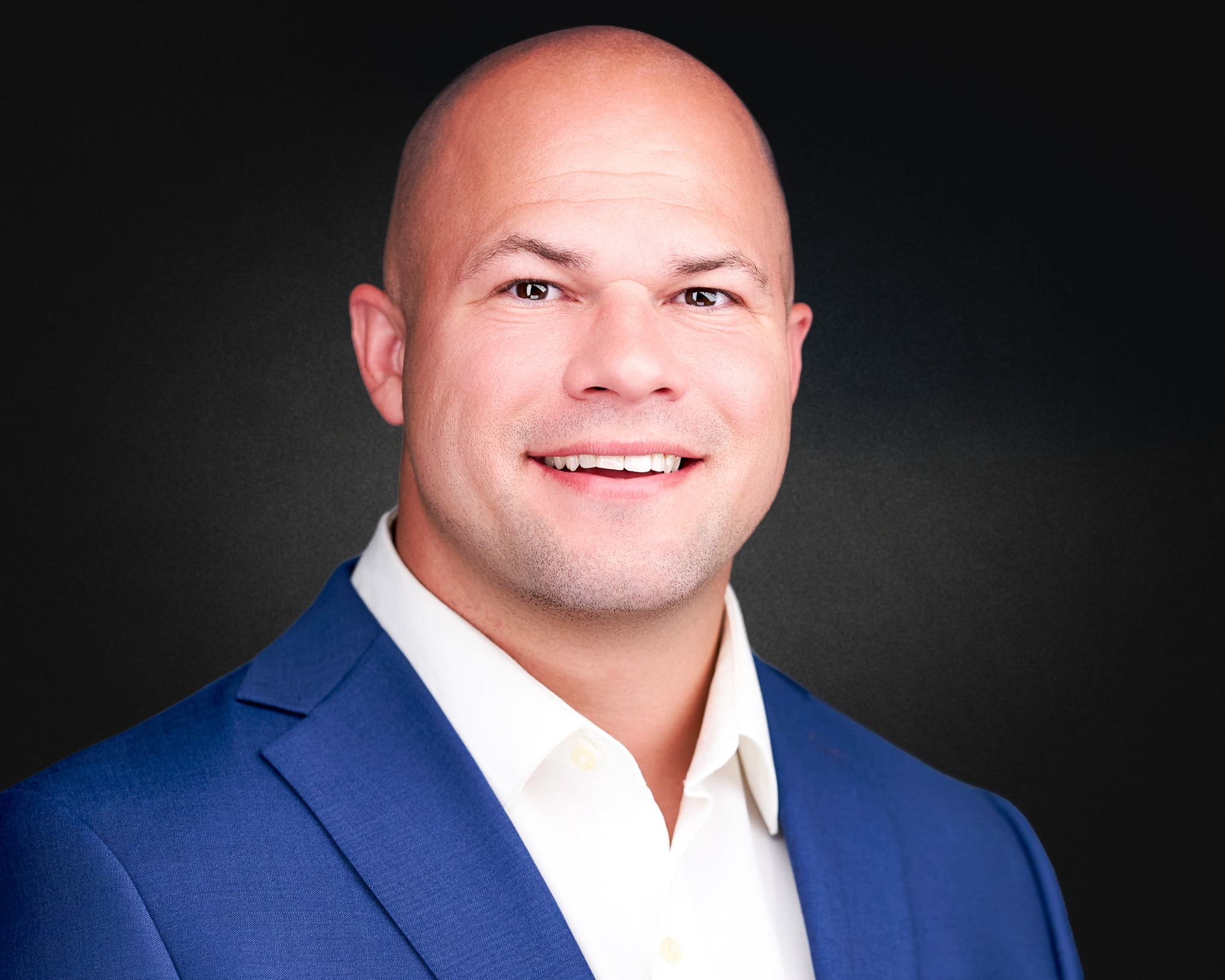 Smiling bald man in a blue blazer and white shirt, studio headshot against a blue backdrop
