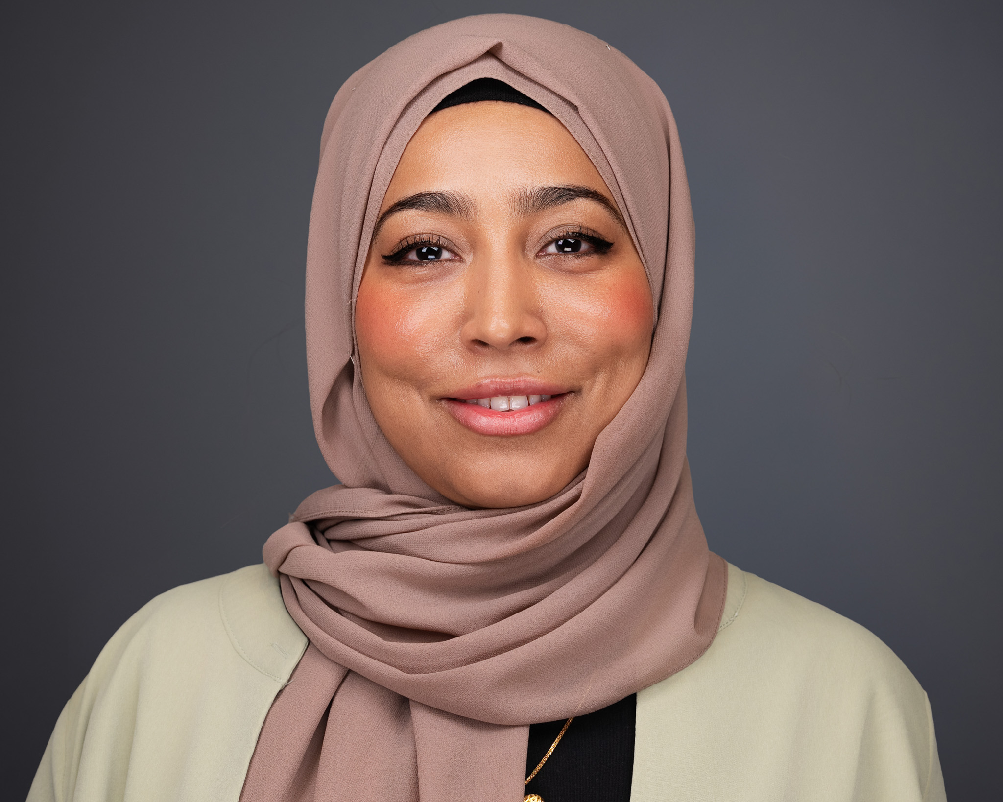 Smiling woman in a beige hijab and cream blazer, studio headshot against a gray backdrop