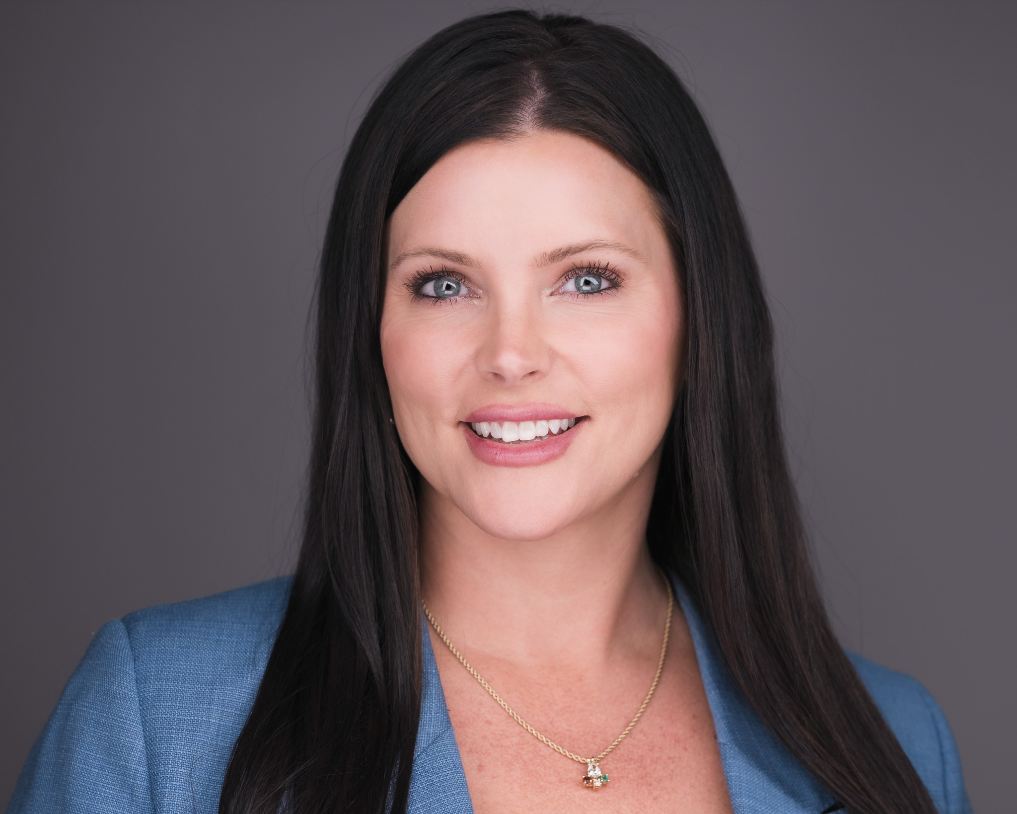 Woman with long dark hair in a blue top, studio headshot against a dark gray backdrop