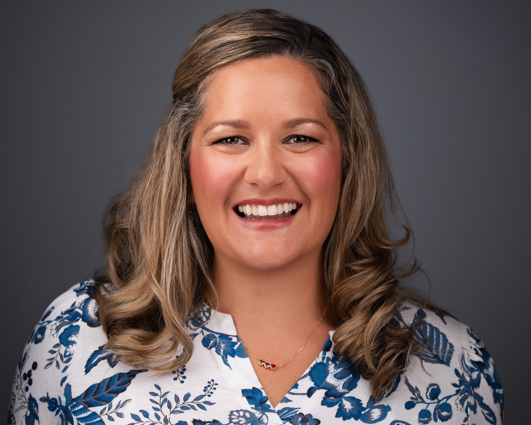 Smiling woman with wavy hair in a floral top, studio headshot against a dark gray backdrop