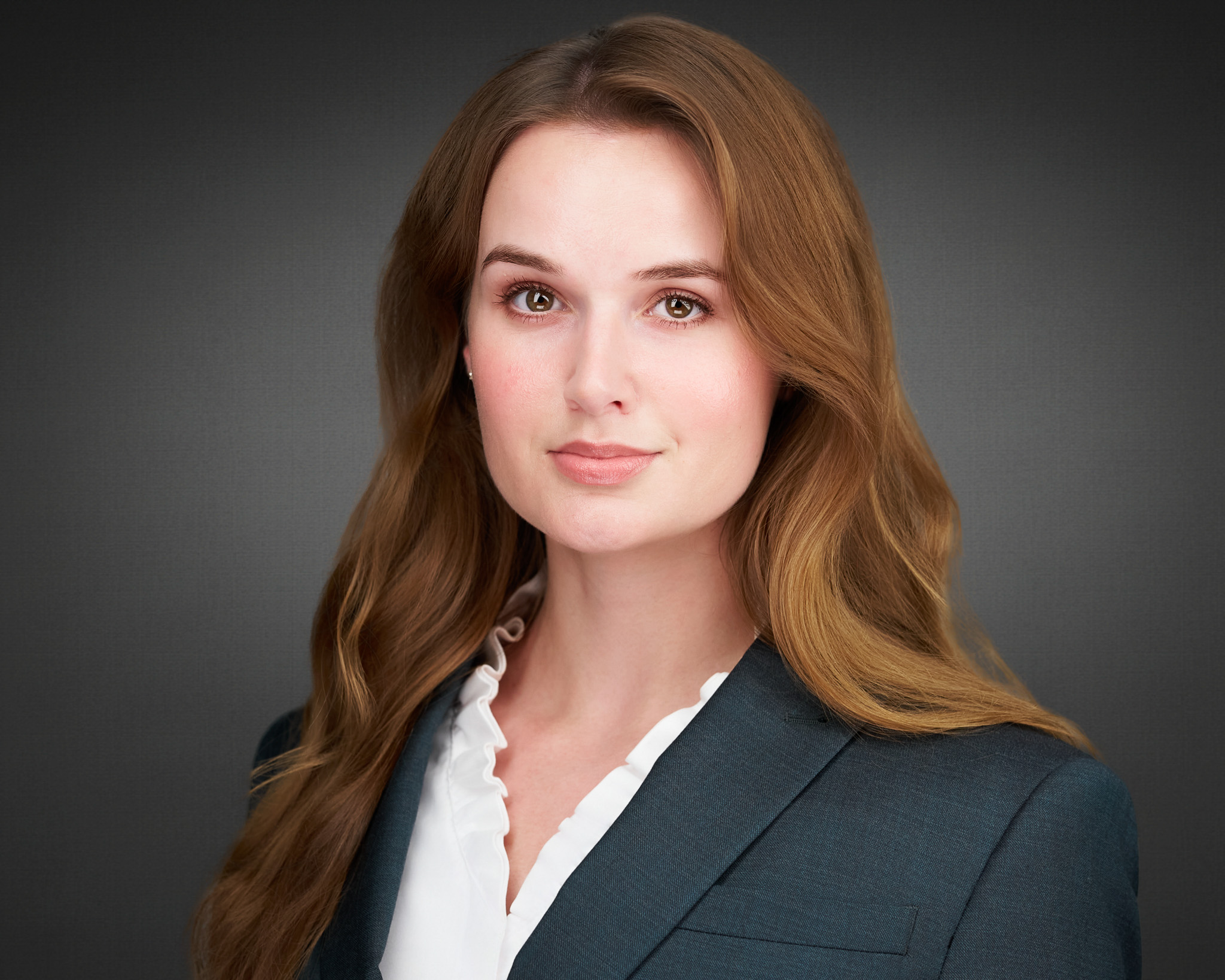 Woman with long auburn hair in a dark blazer, studio headshot against a dark gray backdrop