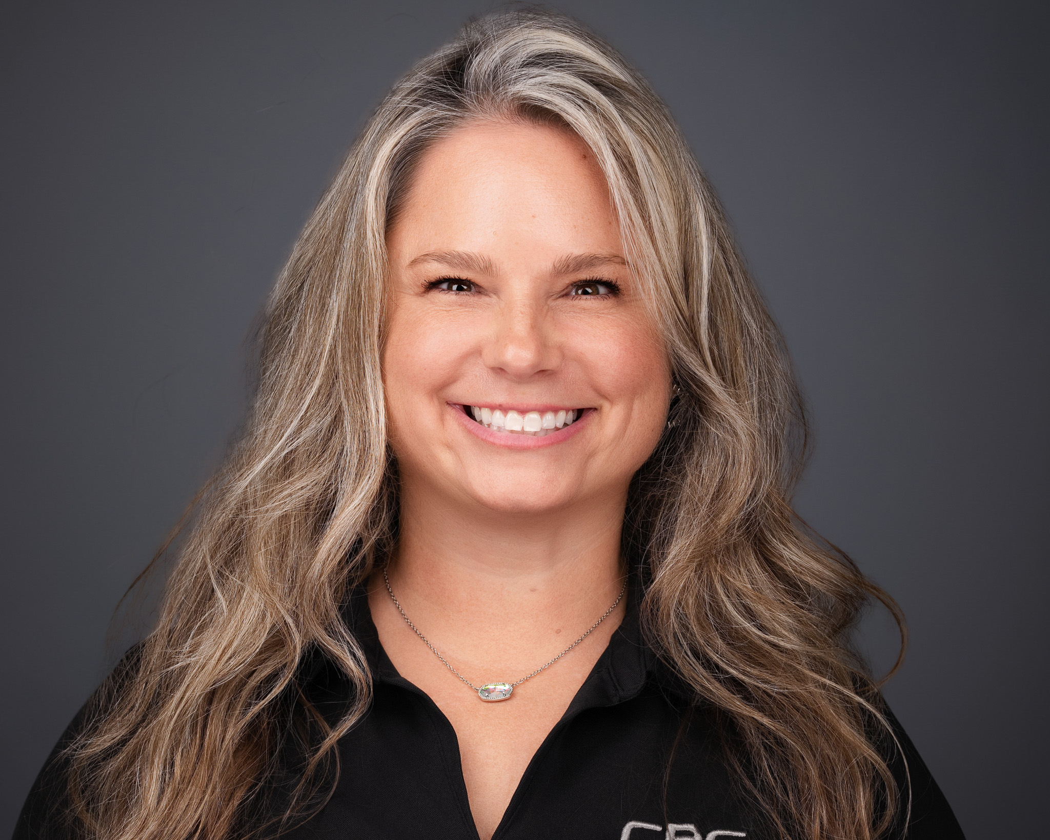 Smiling woman with long wavy blonde hair in a dark top, studio headshot against a dark gray backdrop
