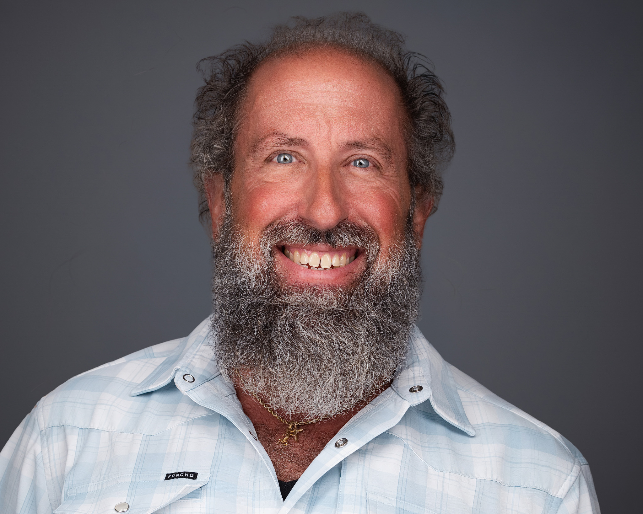 Smiling man with a gray beard and curly hair in a light shirt, studio headshot against a dark gray backdrop