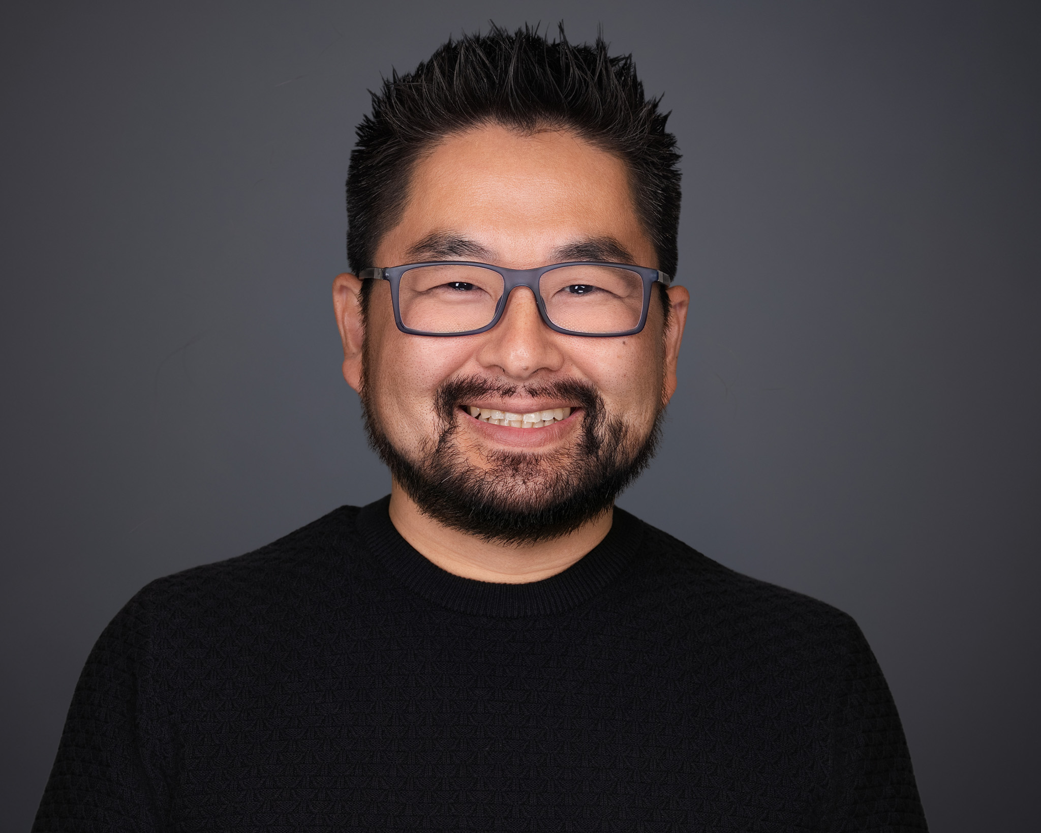 Smiling man with glasses and a beard in a black shirt, studio headshot against a dark gray backdrop
