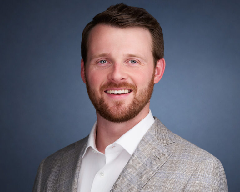 Professional headshot of a smiling man with a short beard wearing a gray plaid blazer and white shirt against a blue-gray background