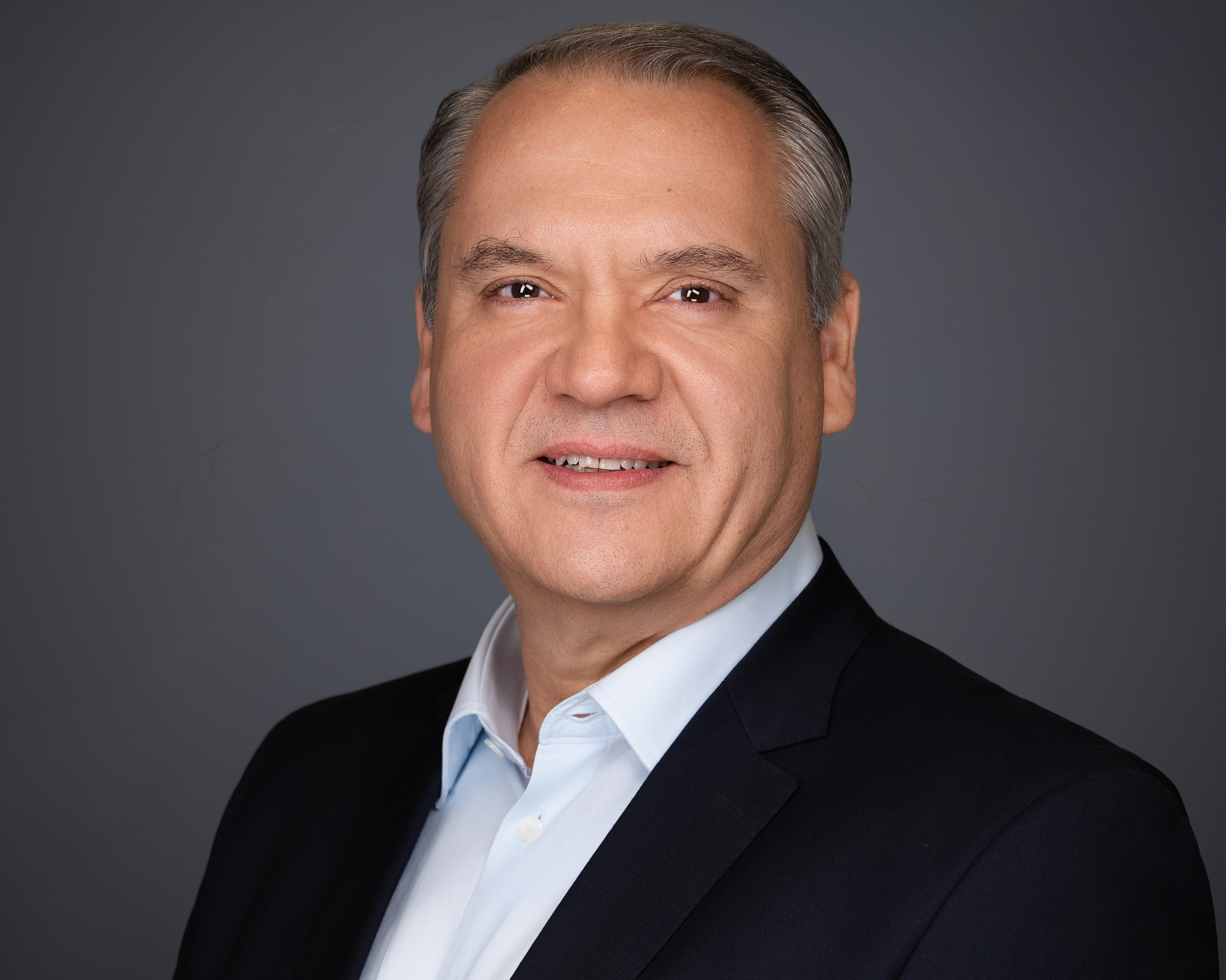 Smiling man with gray hair in a dark blazer, studio headshot against a dark gray backdrop