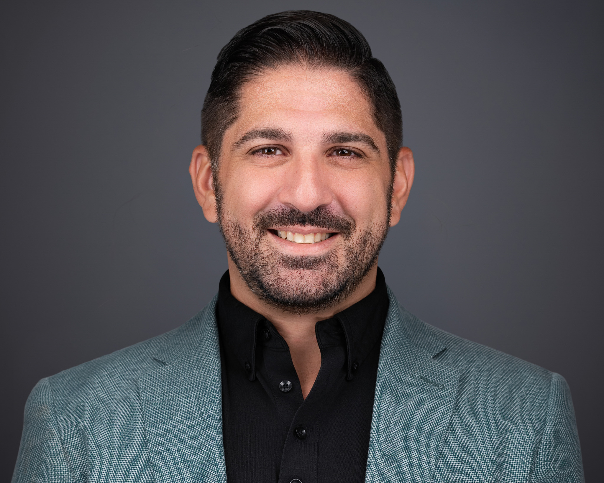 Smiling man with dark styled hair in a teal blazer over a black shirt, studio headshot against a gray backdrop