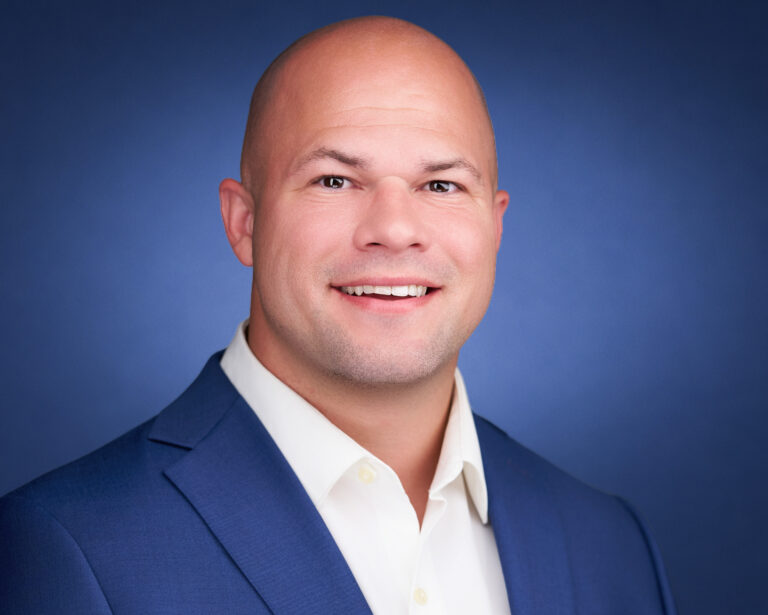 Professional headshot of a smiling bald man in a blue blazer and white shirt against a blue background