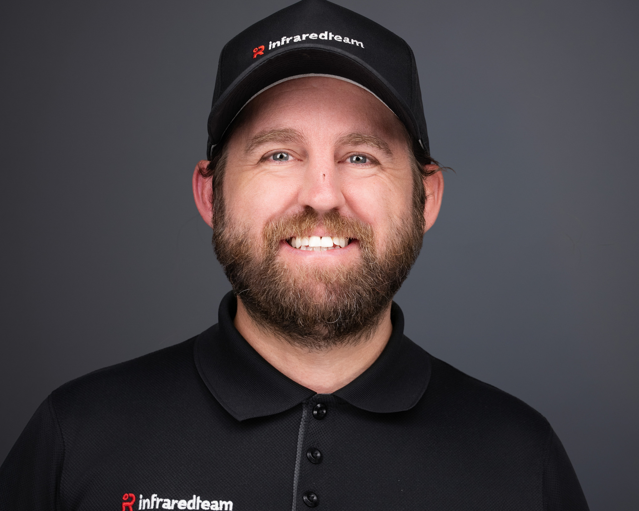 Smiling man with a beard in a black cap and black shirt, studio headshot against a dark gray backdrop