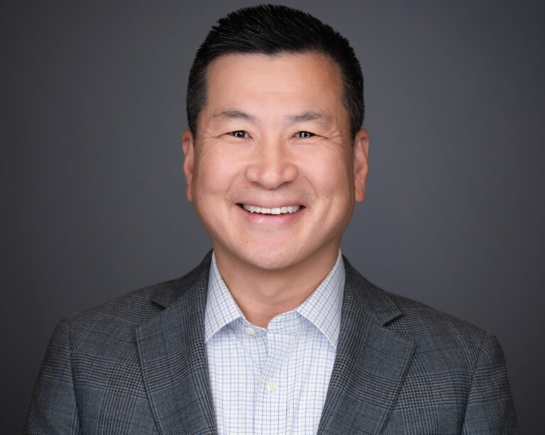 Professional headshot of a smiling man with short dark hair wearing a gray blazer over a light blue shirt against a gray background