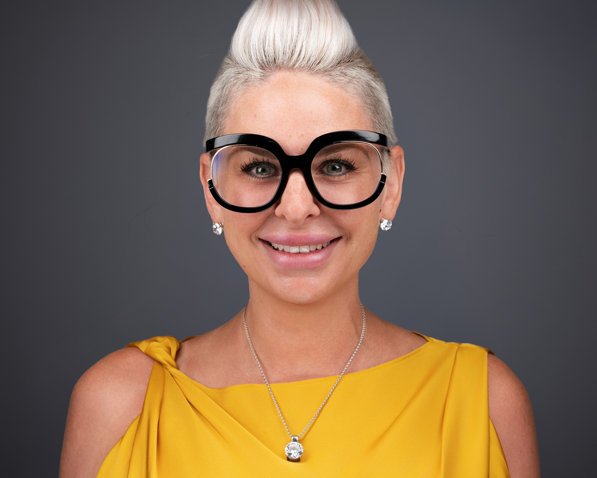 Smiling woman with white hair in a bun, bold glasses and a yellow top, studio headshot against a dark gray backdrop