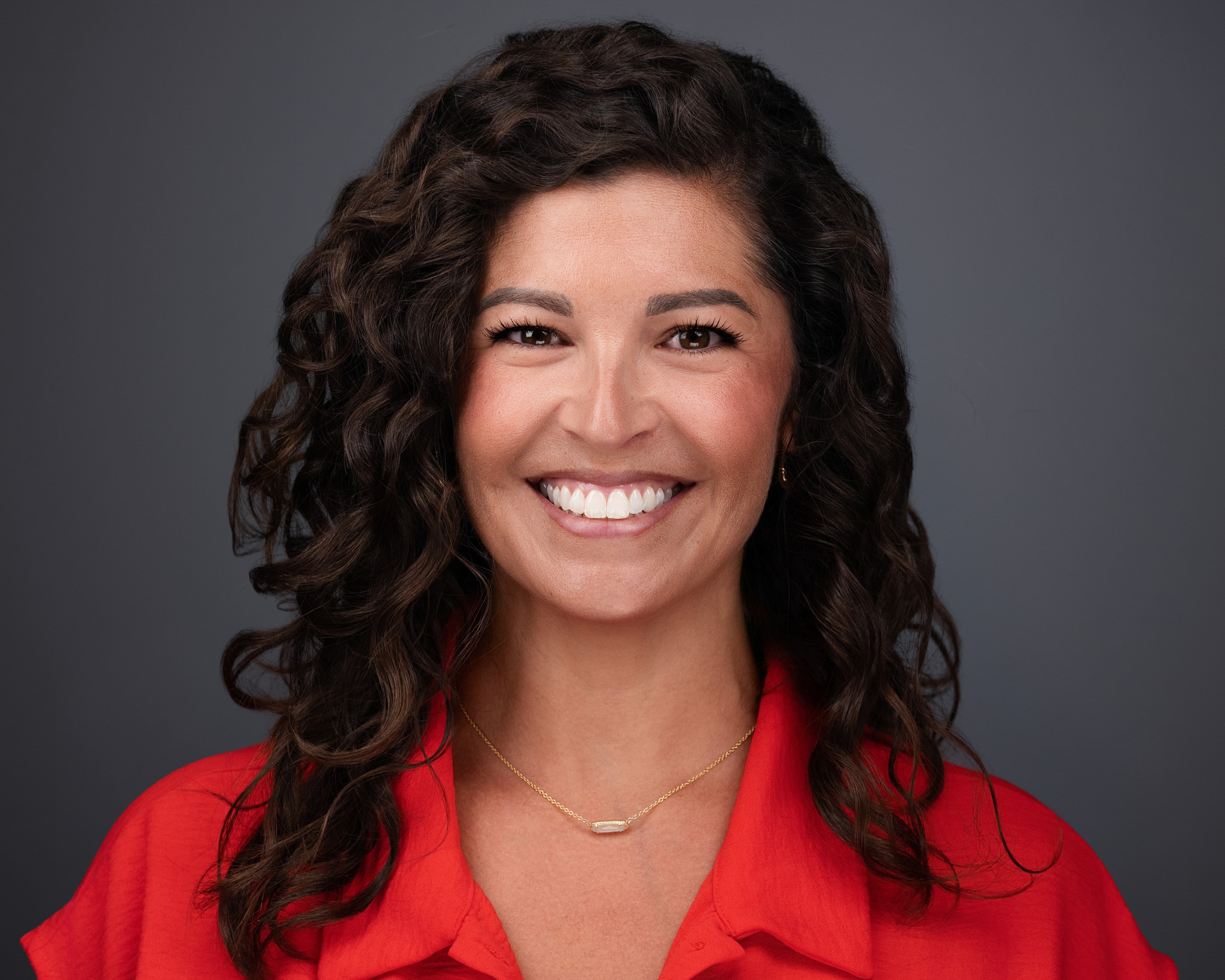 Smiling woman with dark curly hair in a red top, studio headshot against a dark gray backdrop
