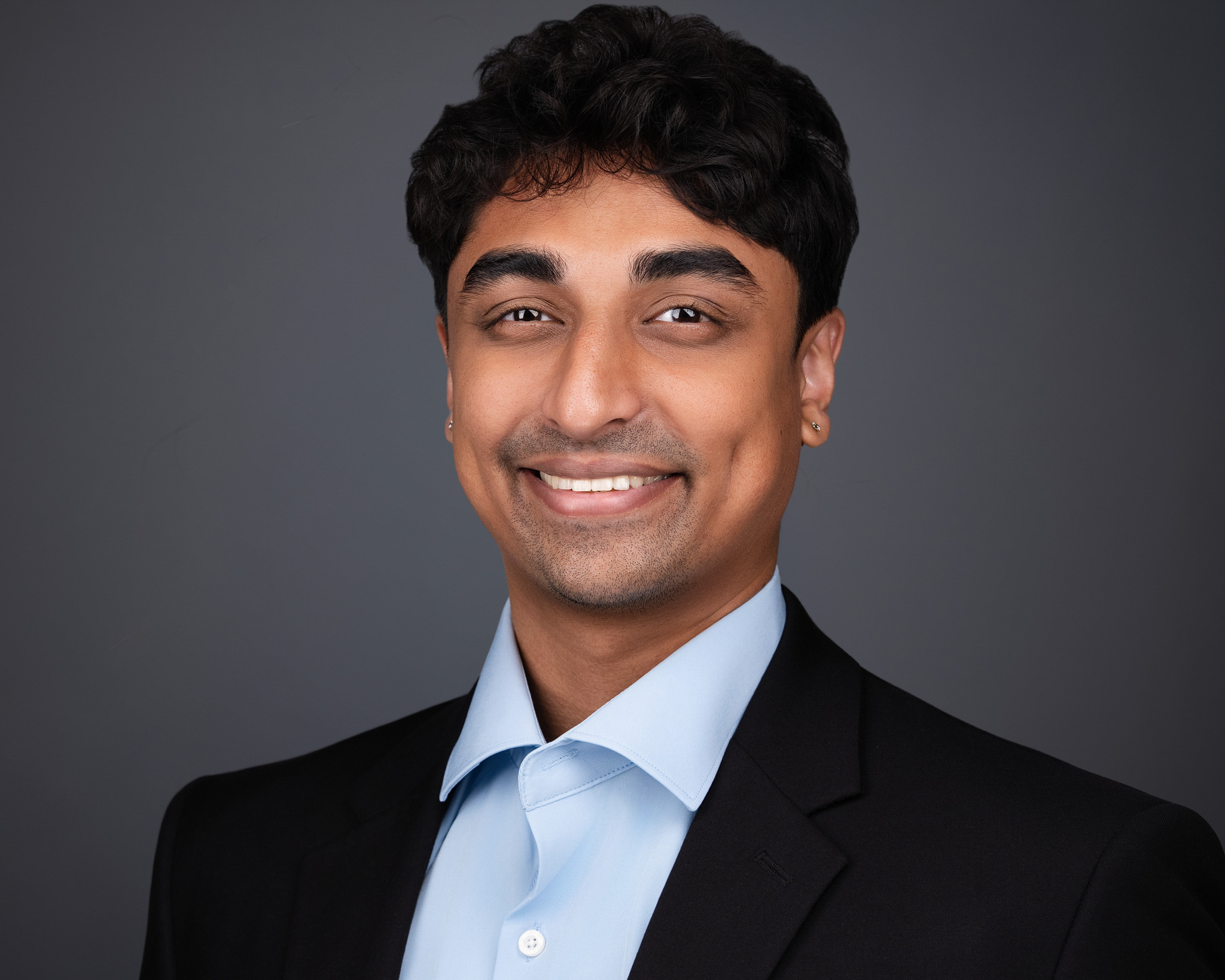 Smiling man in a navy blazer and light blue shirt, studio headshot against a dark gray backdrop