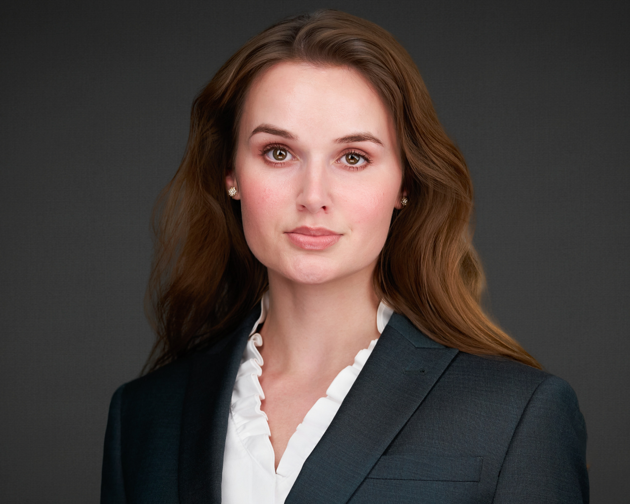 Woman with long auburn hair in a charcoal blazer and white blouse, studio headshot against a dark gray backdrop