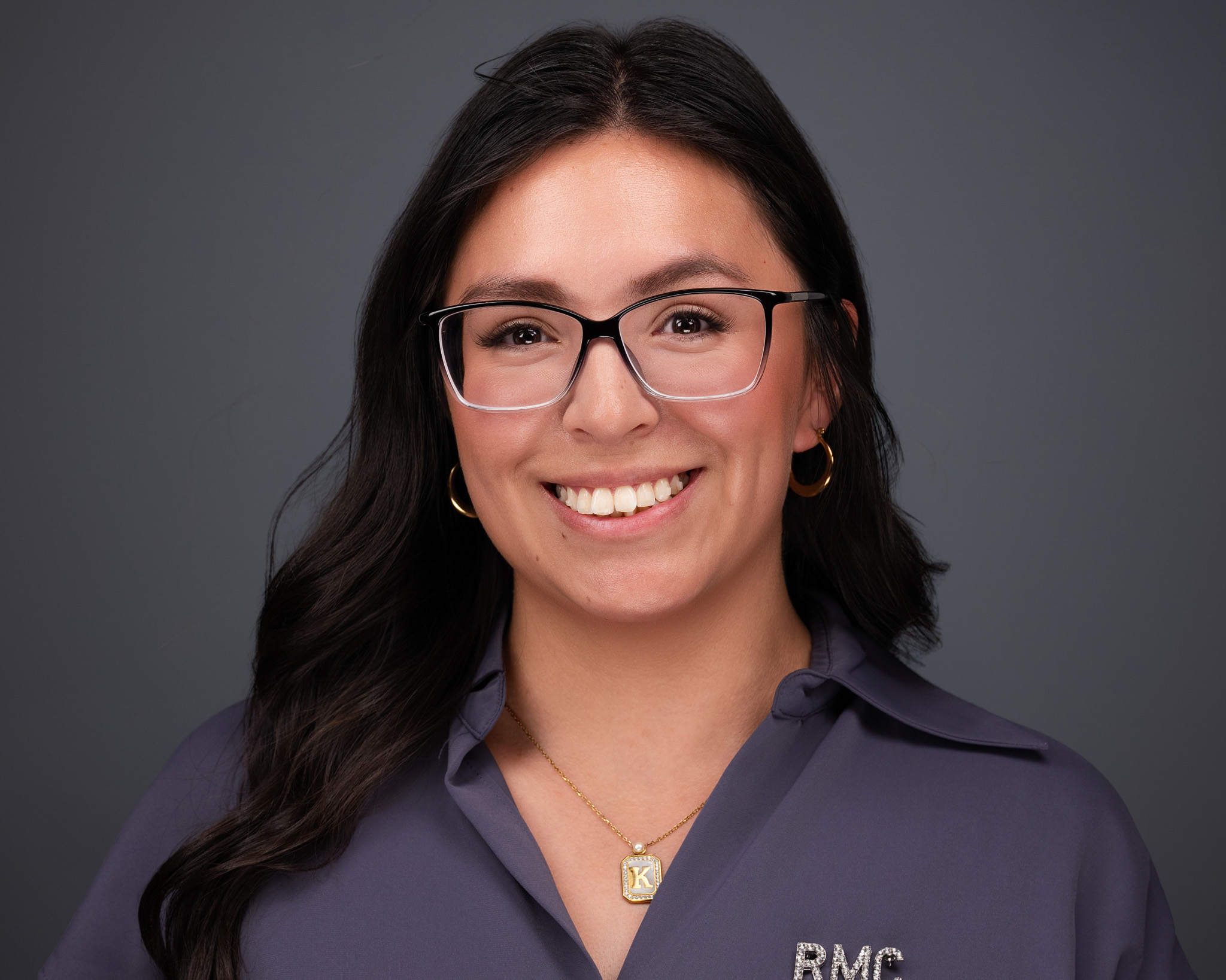 Smiling woman with long dark hair and glasses in a gray top, studio headshot against a dark gray backdrop