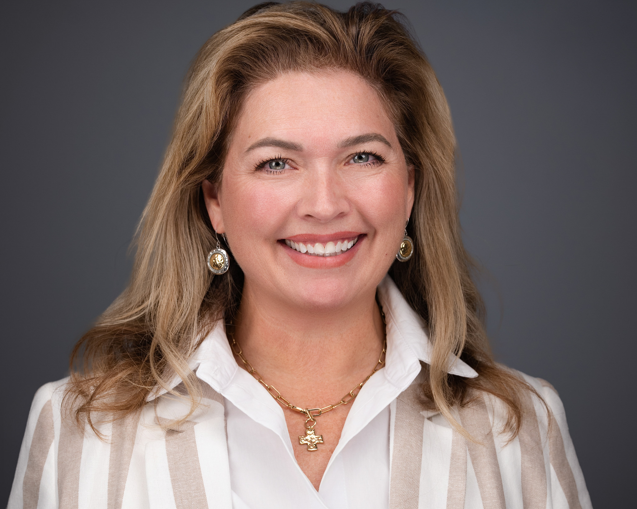 Smiling woman with wavy blonde hair in a striped blouse, studio headshot against a dark gray backdrop