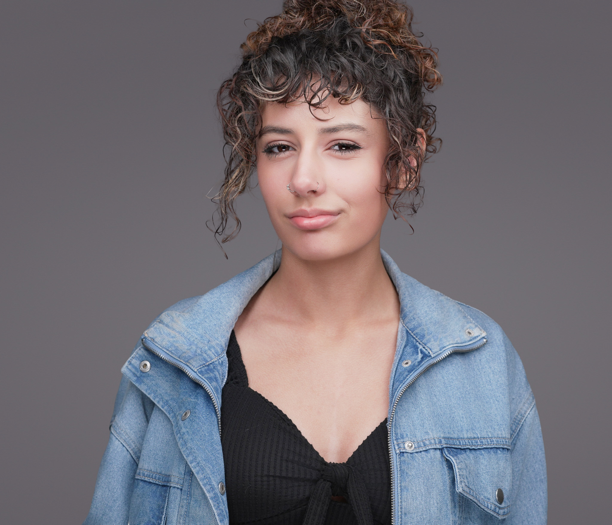Smiling woman with short curly hair in a denim jacket, studio headshot against a dark gray backdrop