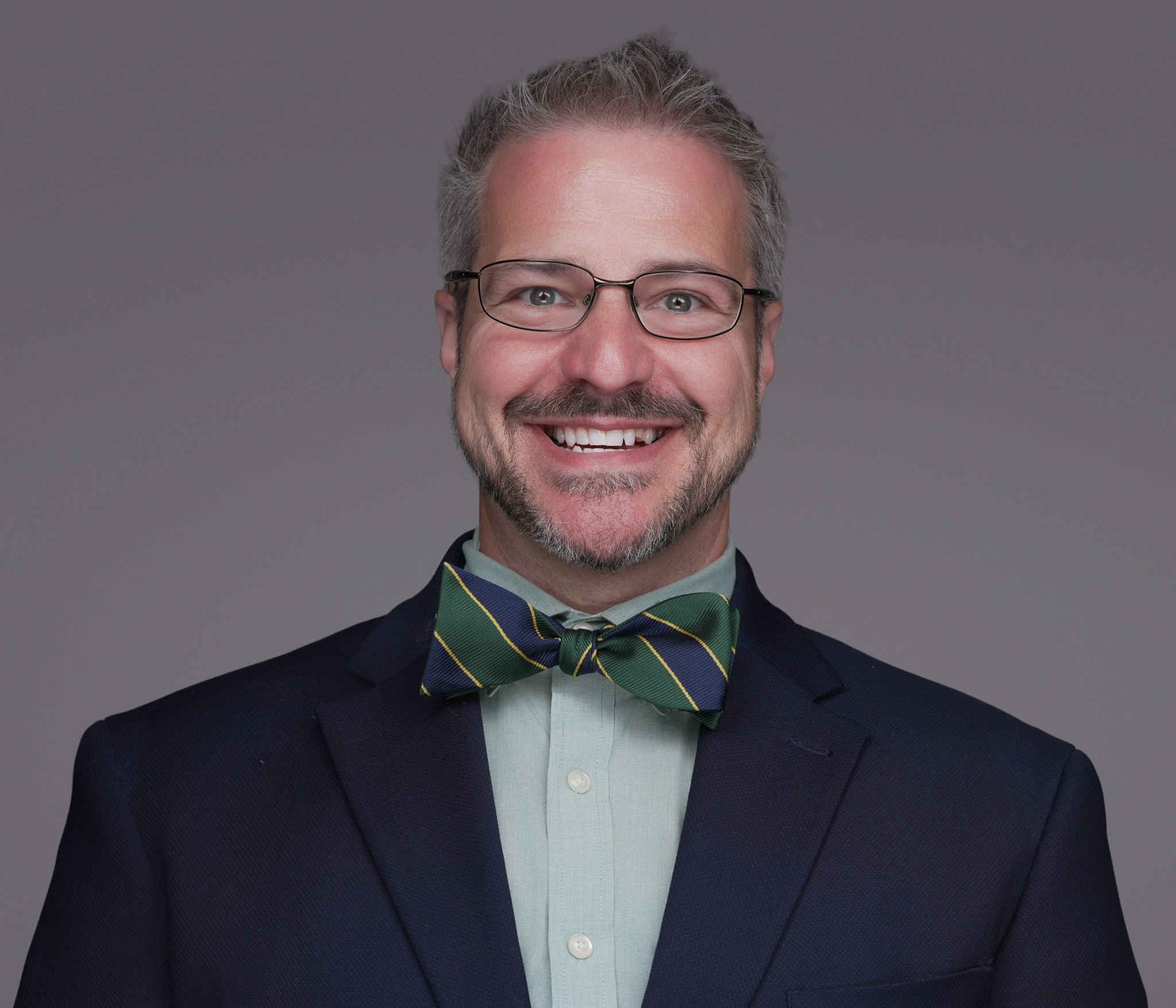 Smiling man with glasses in a navy blazer and green bow tie, studio headshot against a gray backdrop