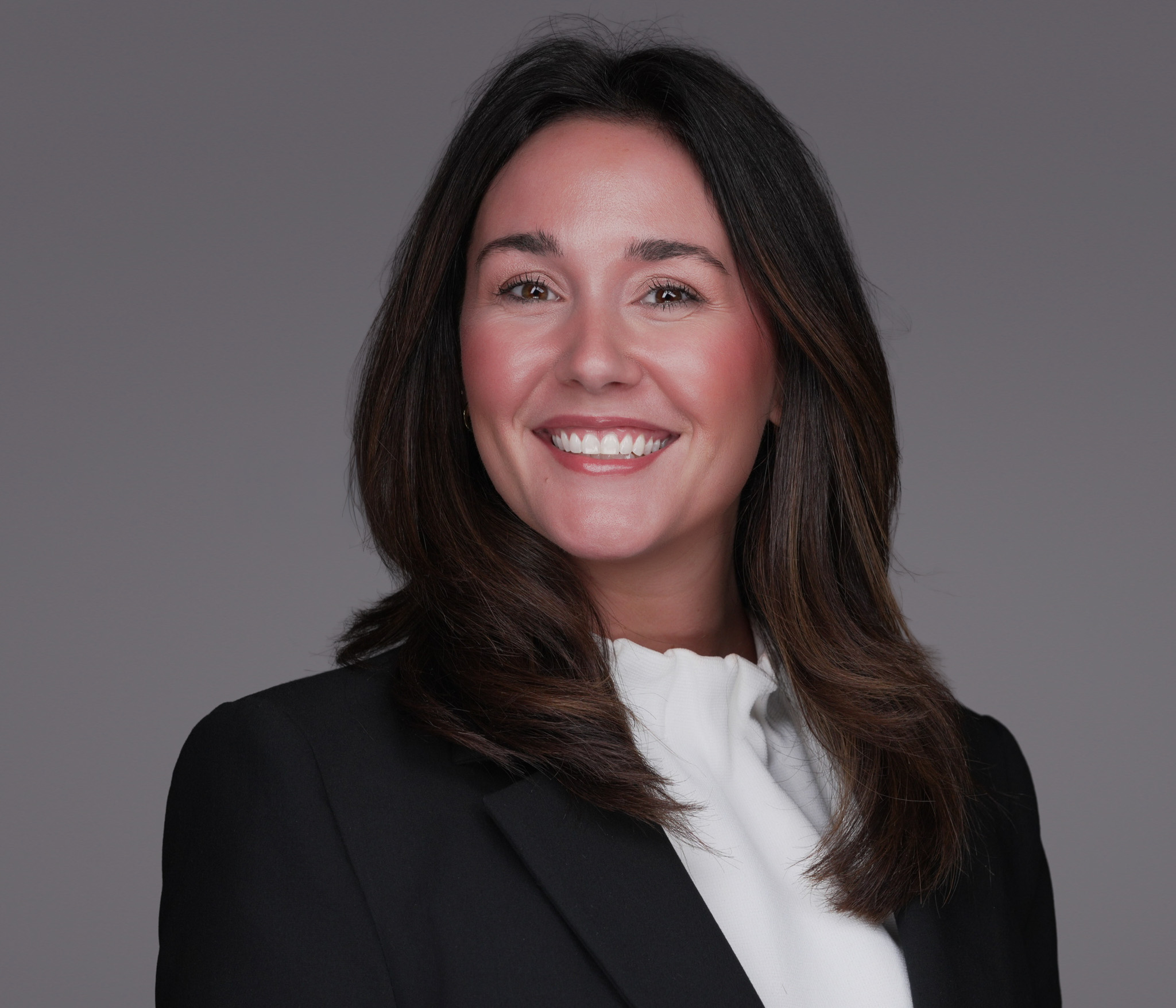 Smiling woman with long dark hair in a black blazer, studio headshot against a dark gray backdrop