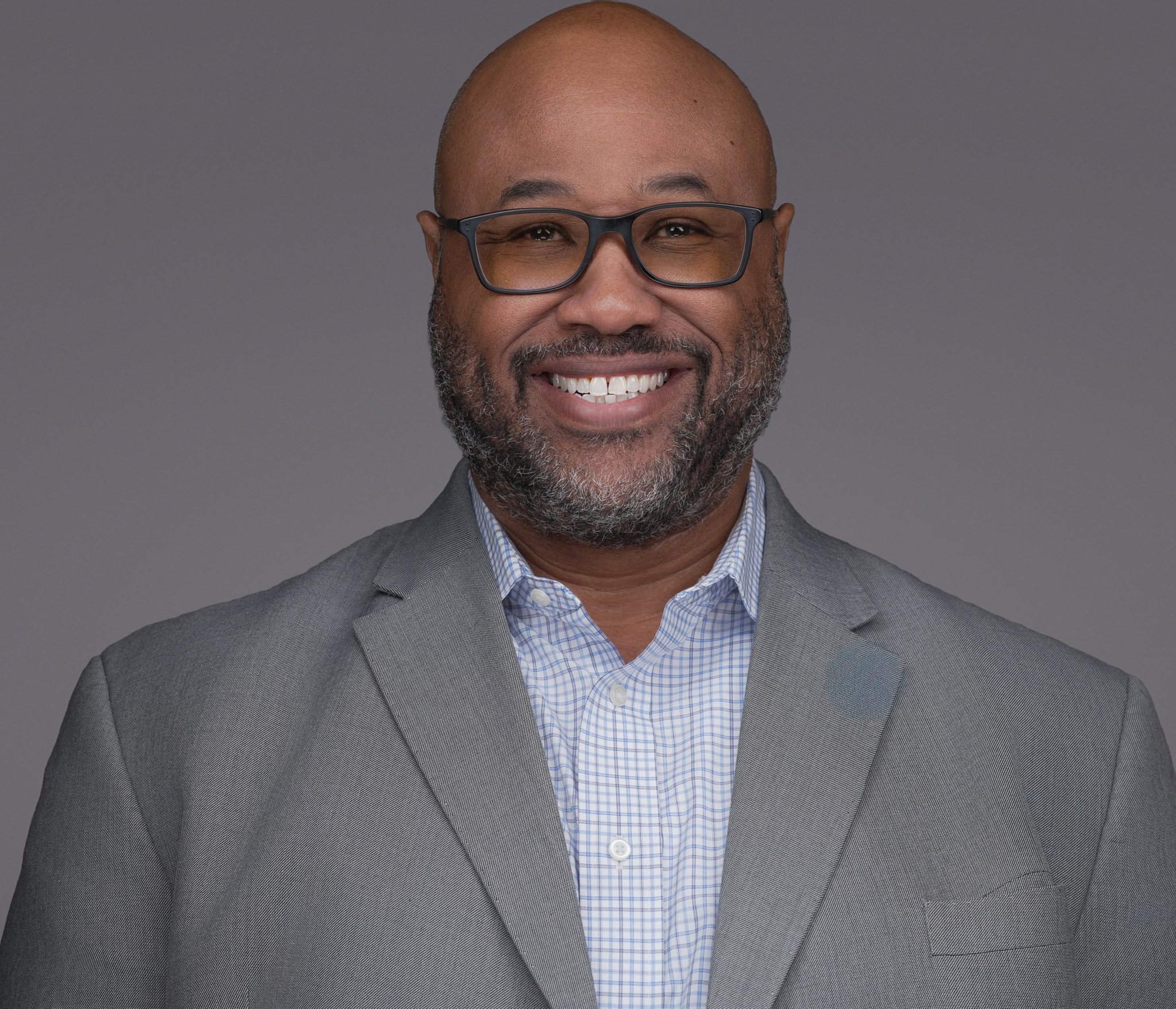 Smiling bald man with glasses in a gray blazer, studio headshot against a dark gray backdrop